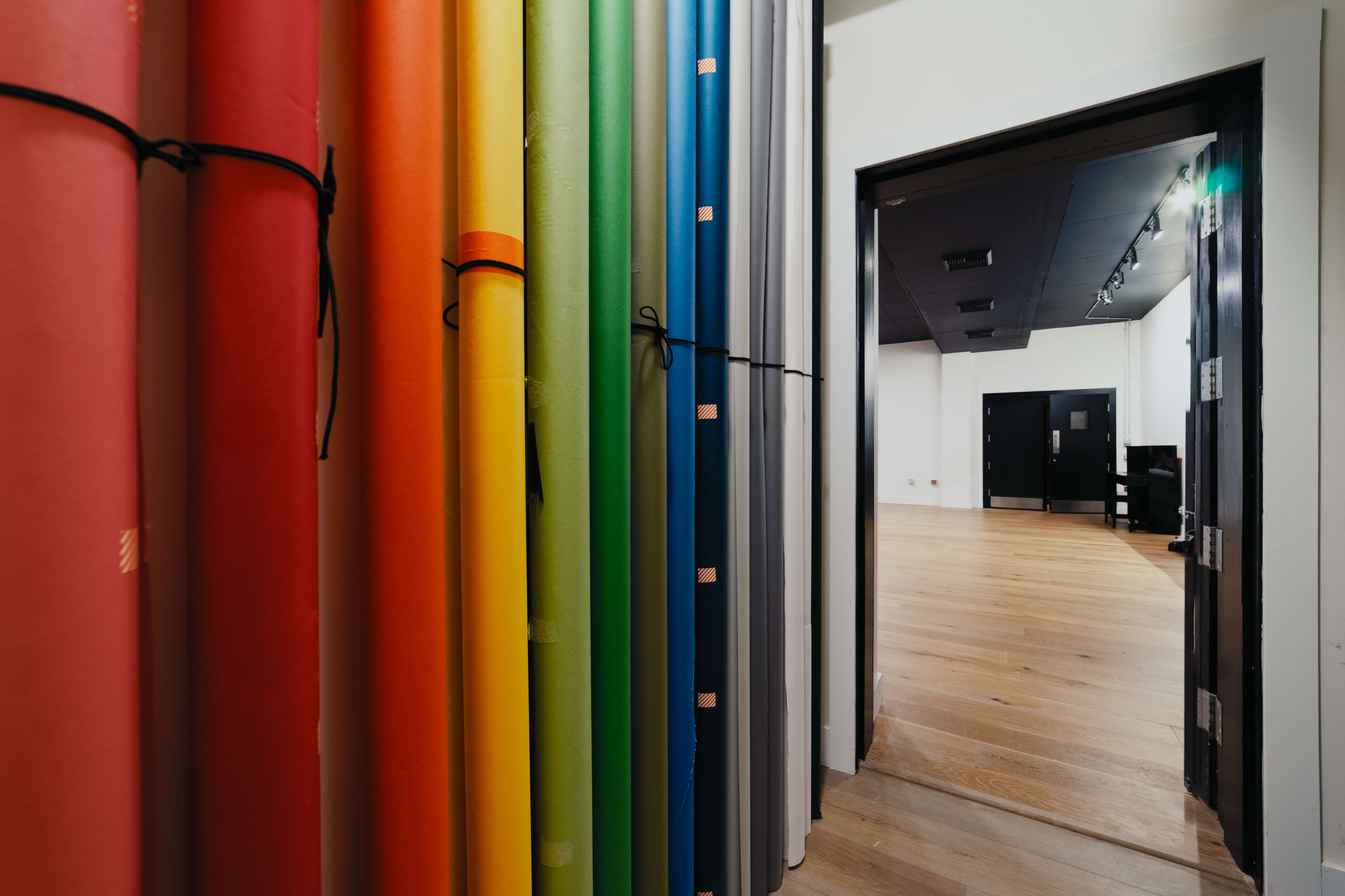 The image displays a collection of rolled paper in various colors leaning against a wall, with an open doorway leading into a room with a wooden floor.