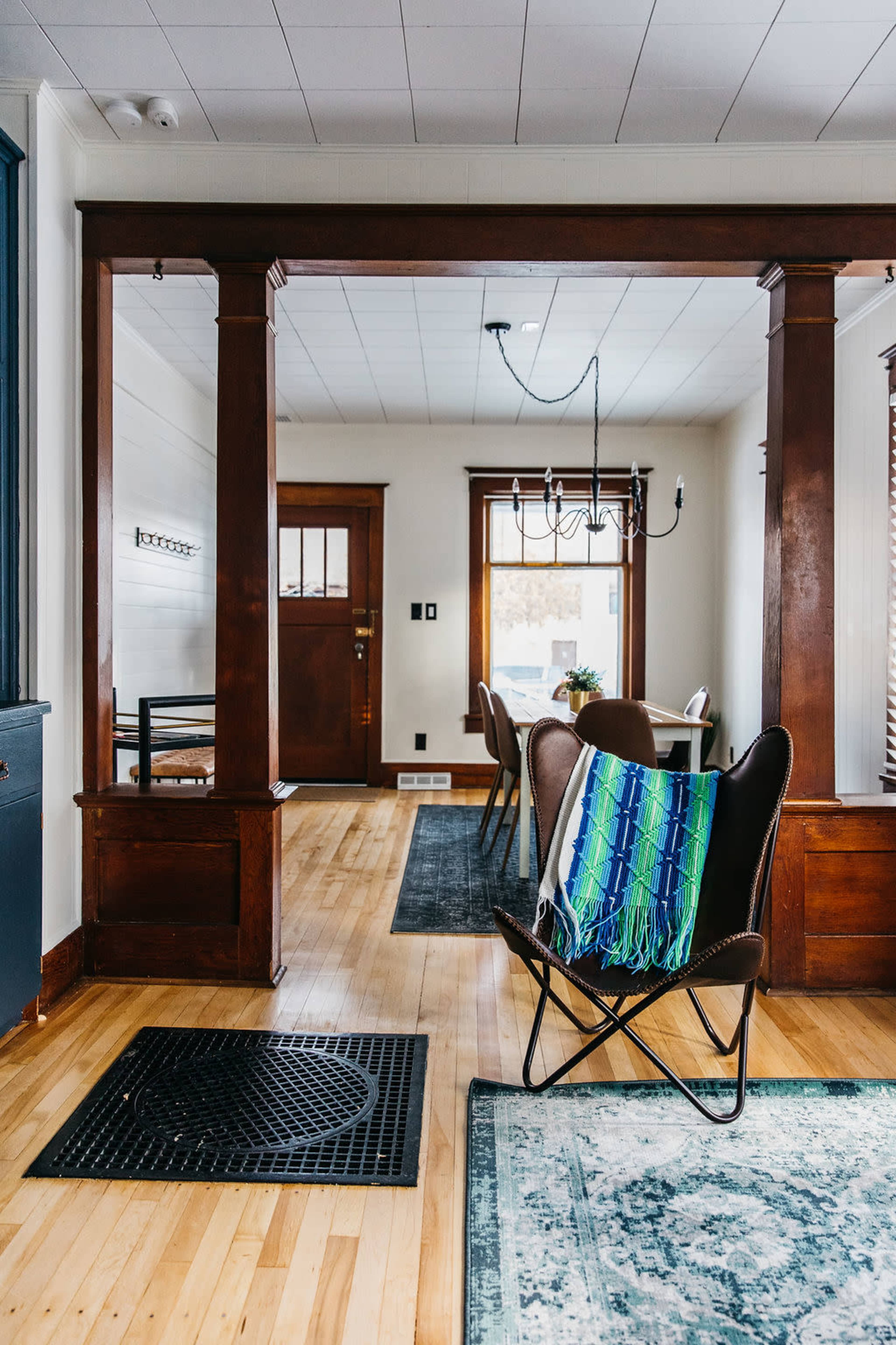 A spacious interior featuring a wooden entrance with a door, a dining area with a chandelier, and a chair draped with a colorful blanket.