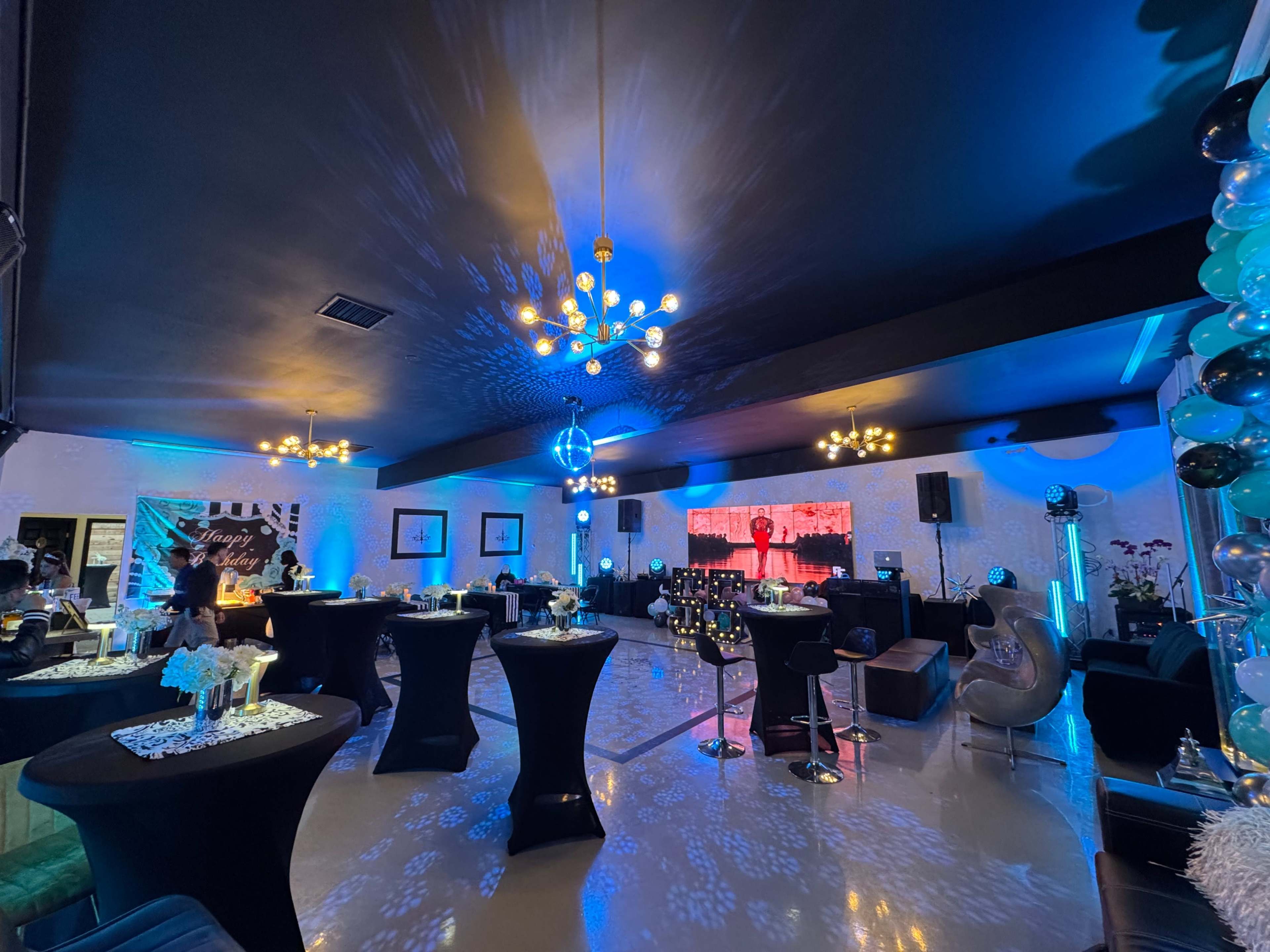 A spacious event venue is decorated with black and blue lighting, featuring tables with centerpieces, lounge seating, and a large screen displaying visuals.