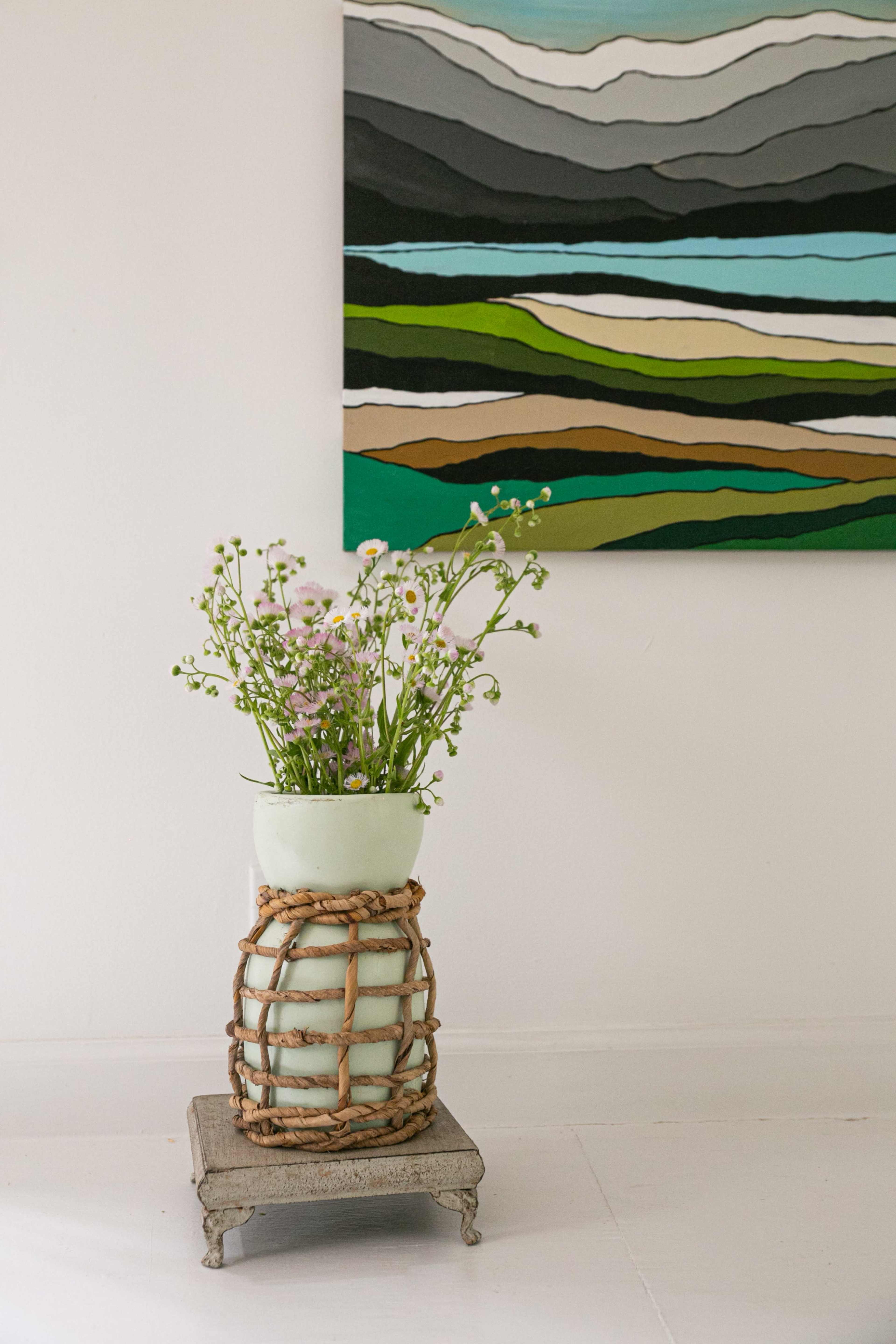 A floral arrangement in a textured vase rests on a small metallic stand in front of a colorful abstract painting.
