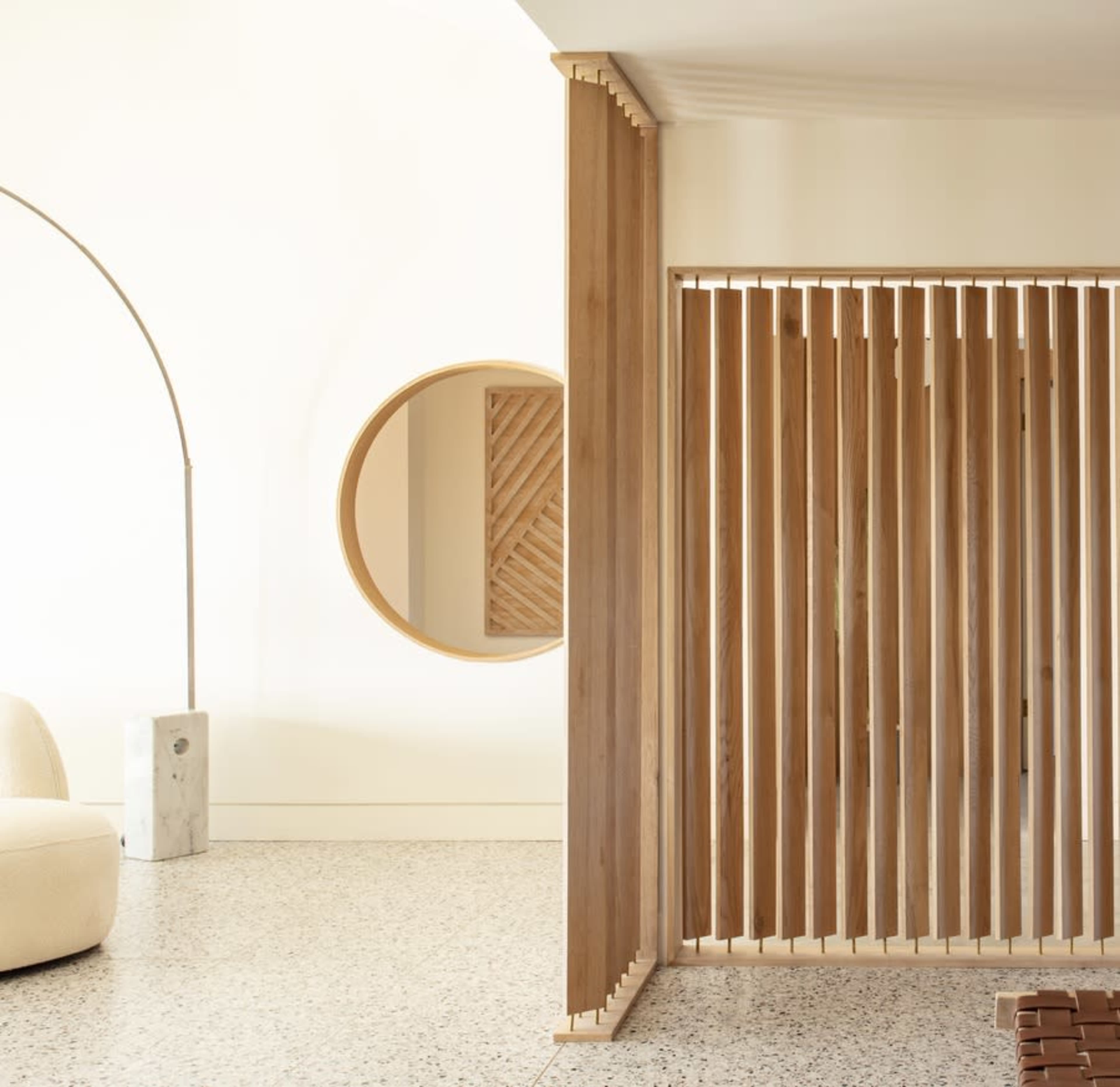 The image showcases a modern interior with wooden slat partitioning, a circular mirror, and a curved lounge chair, all set against a light-colored wall and speckled floor.
