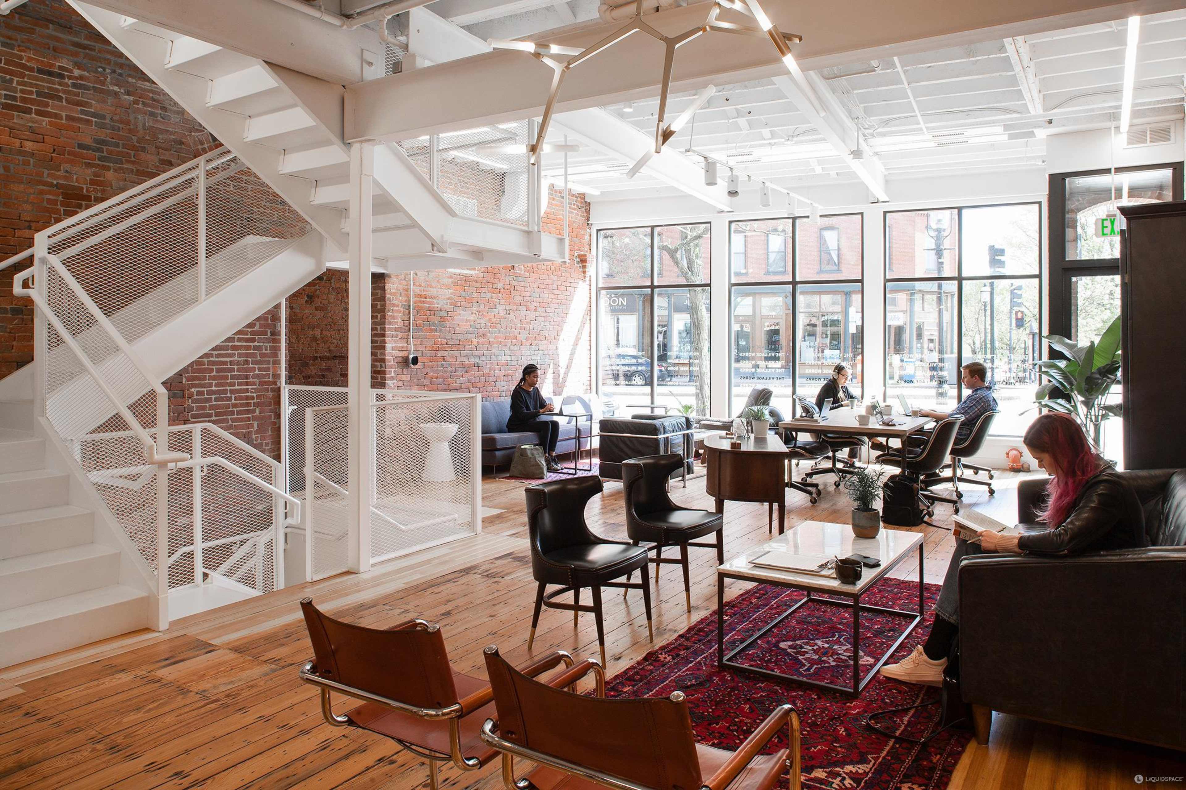 The image shows a modern office space featuring a mix of seating areas, large windows, and a staircase with an exposed brick wall.