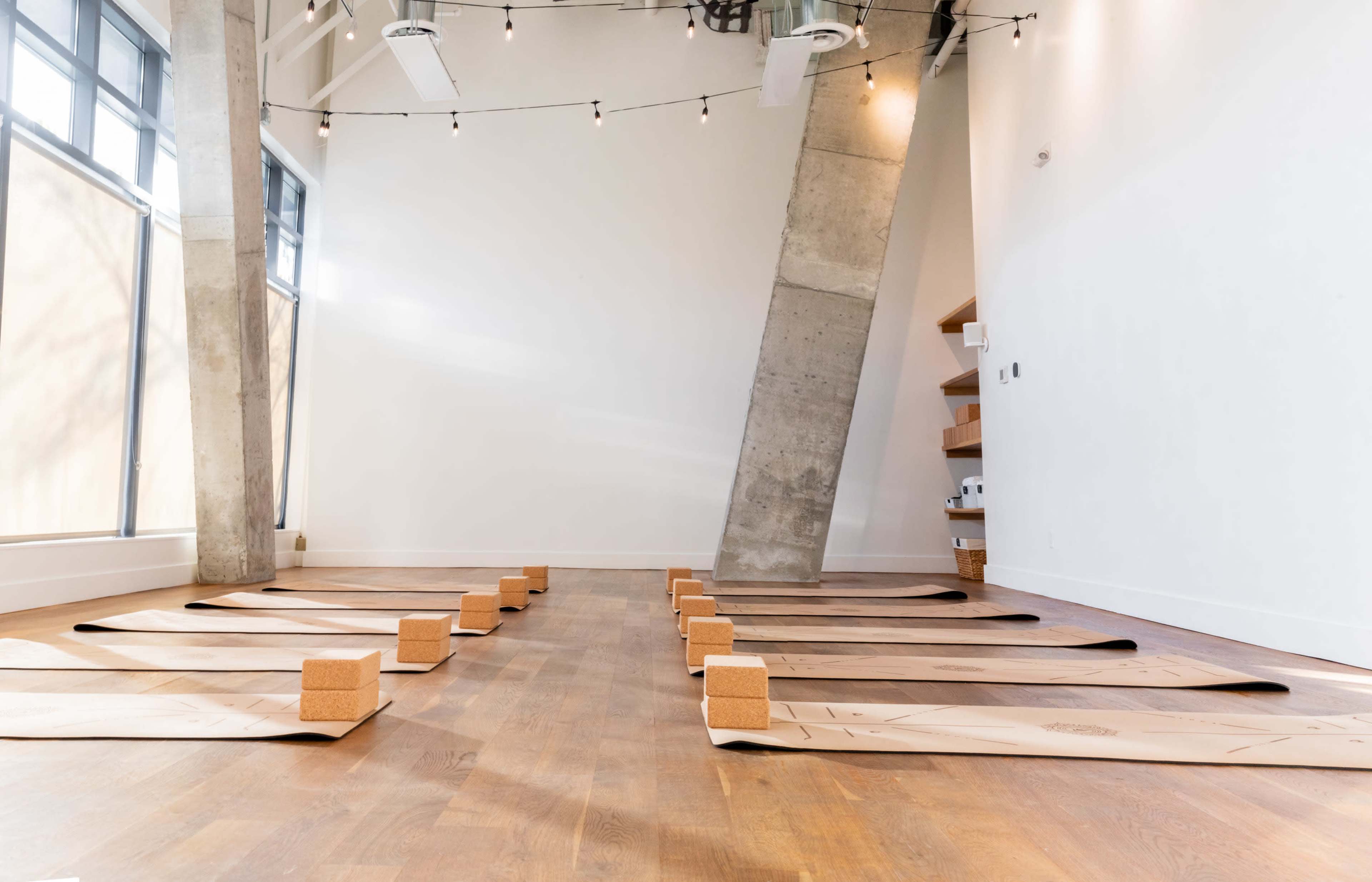 A yoga studio features wooden flooring with neatly arranged mats and cork blocks under soft lighting.