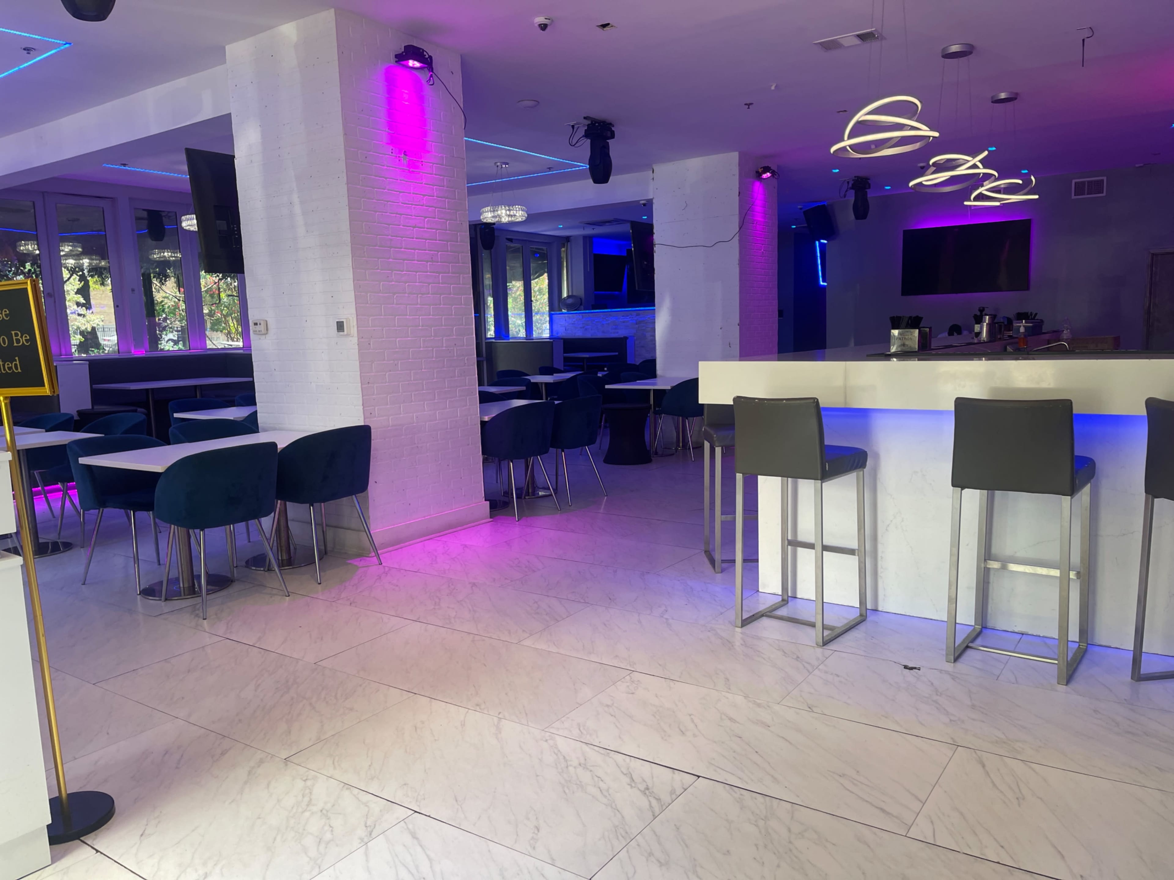 A modern indoor space featuring a bar area with high stools, tables, and purple lighting.