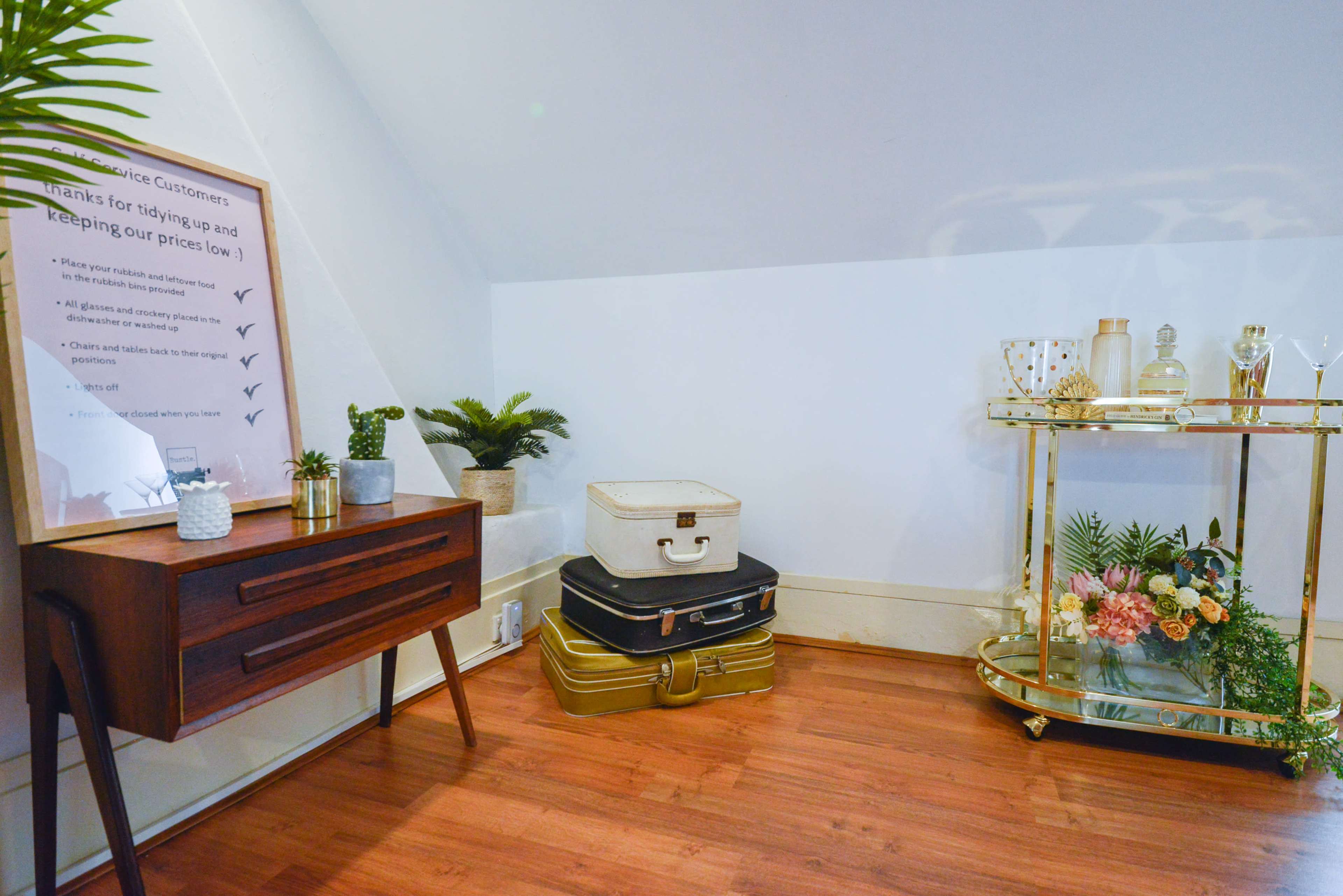 A wooden console table is positioned next to stacked vintage suitcases, with a bar cart displaying decorative items and plants in a well-lit room.
