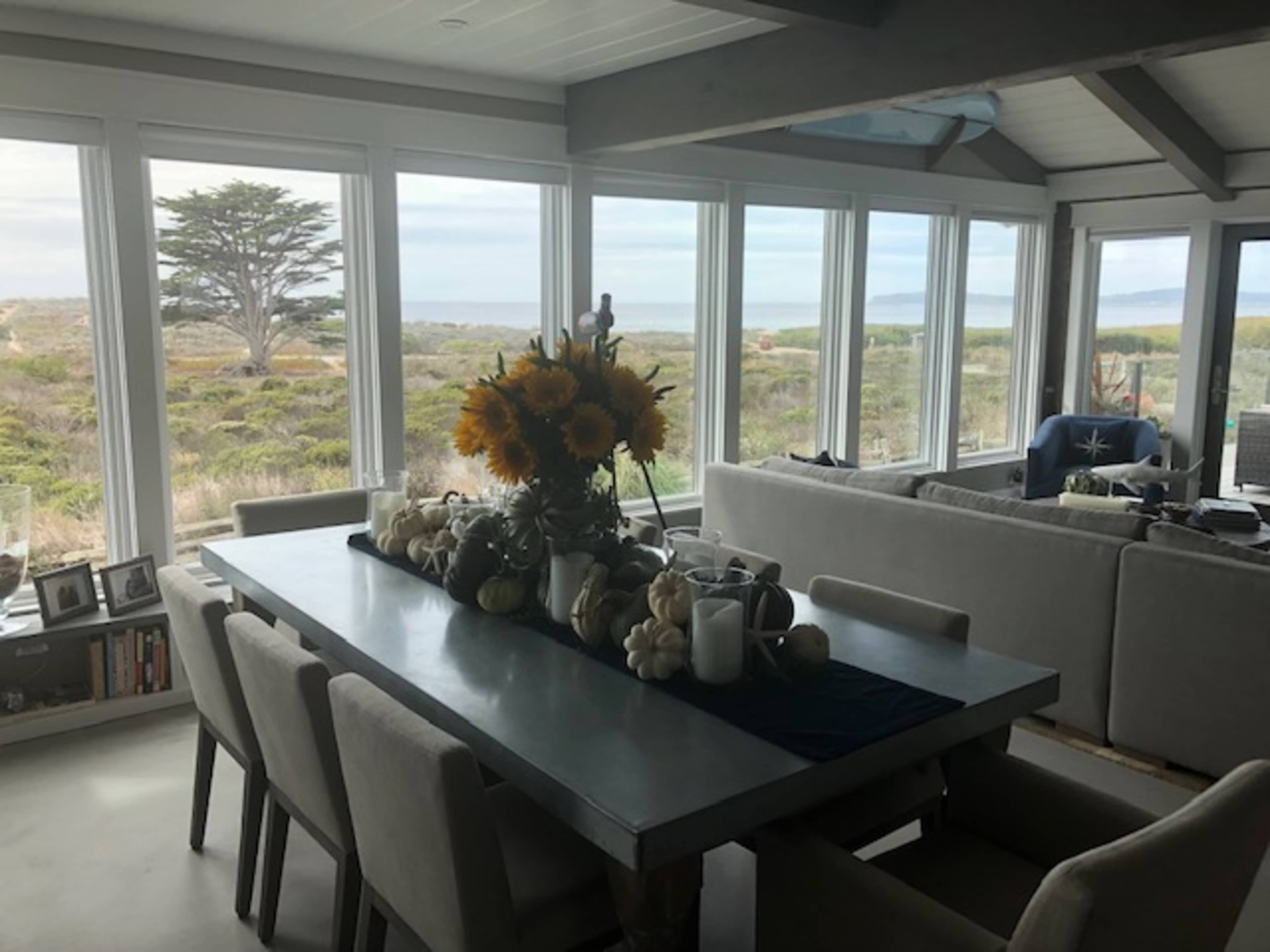 A dining area features a long table adorned with a flower arrangement and decorative items, surrounded by chairs and open windows that showcase a coastal view.