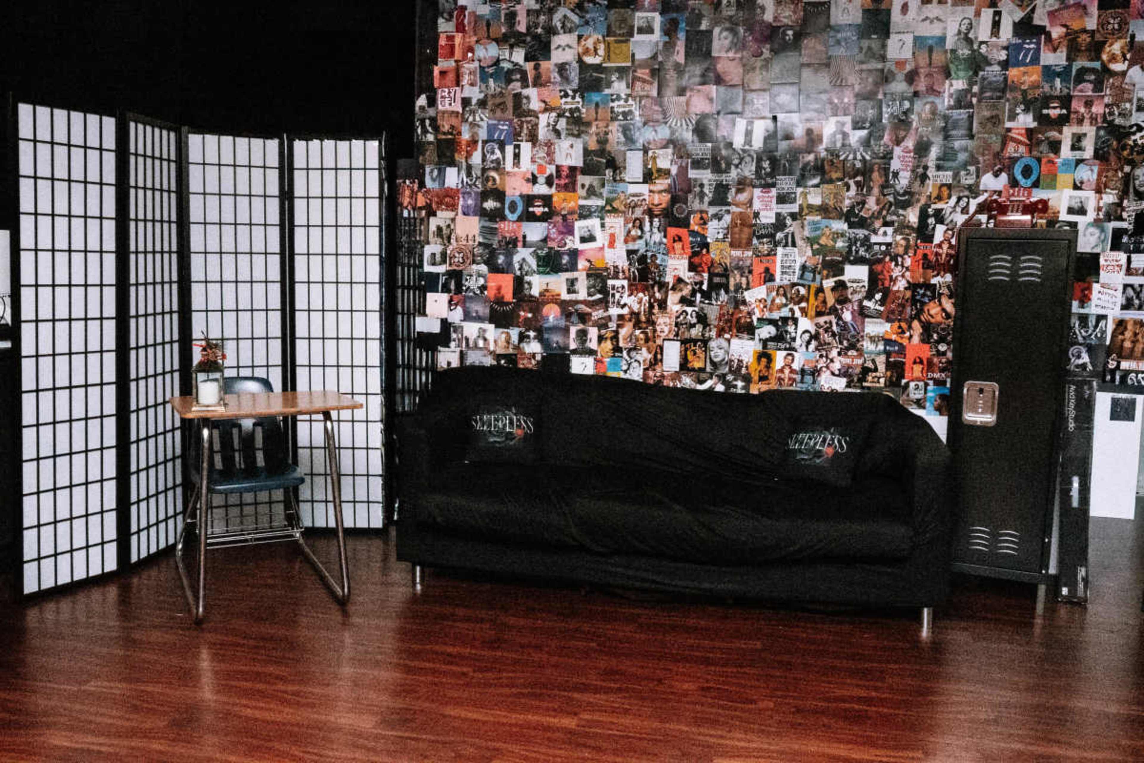 The scene features a black couch facing a wall covered in various posters, with a folding screen on one side and a table with a chair nearby.