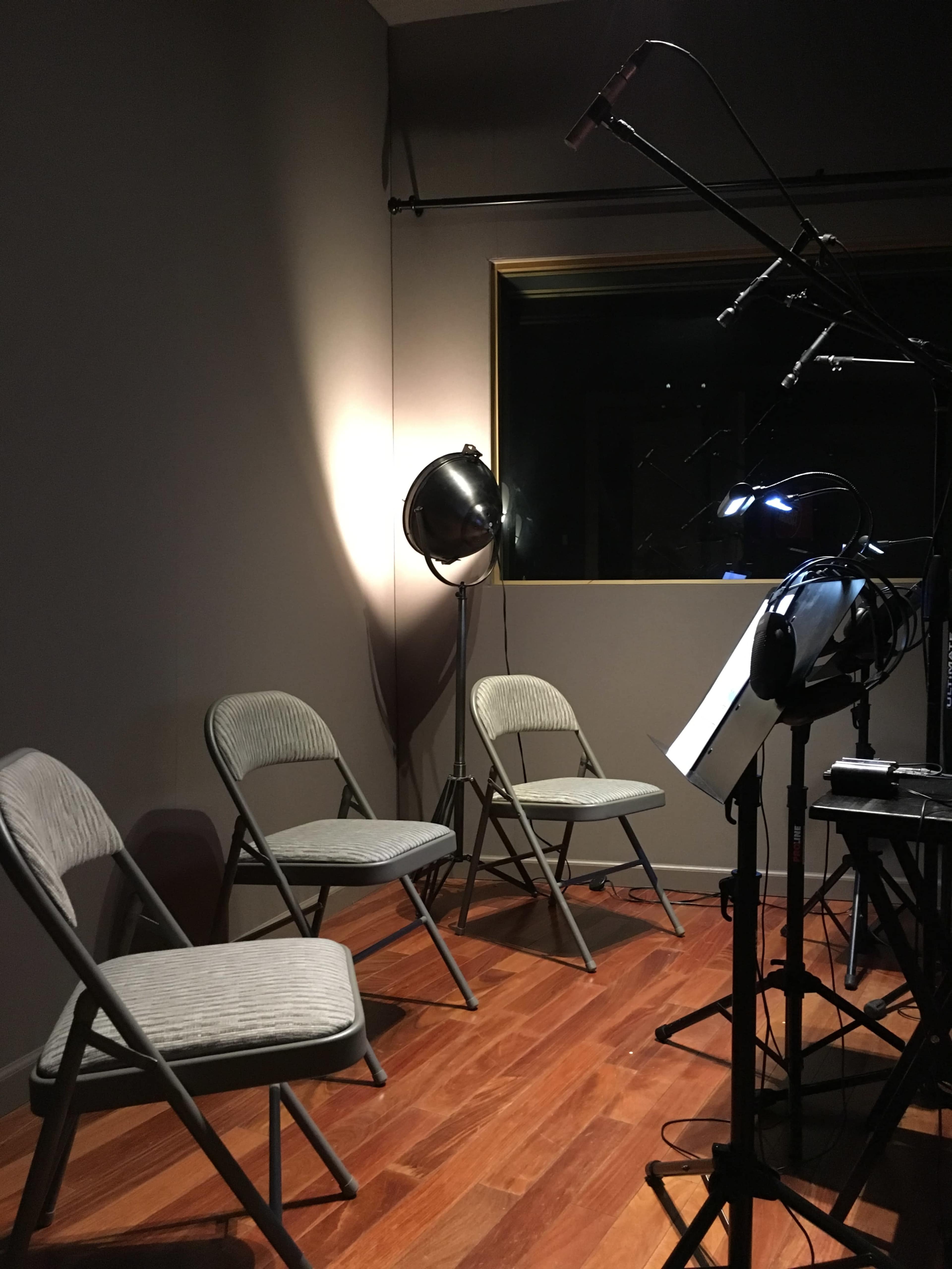 The image shows a small audio recording studio with three empty folding chairs and a standing lamp illuminating the space.
