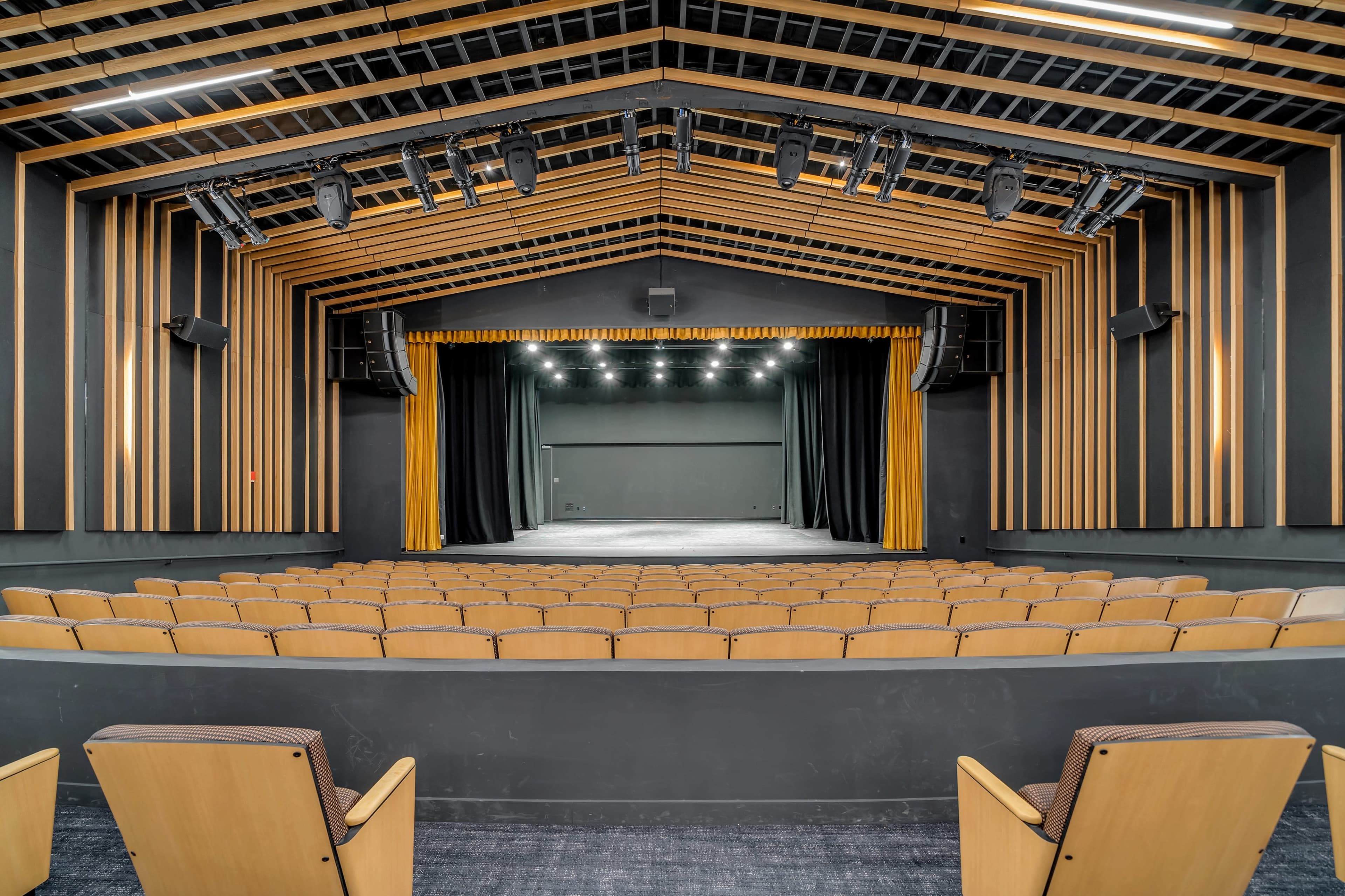An empty auditorium with wooden seating facing a proscenium stage, featuring black curtains and a wooden accent wall.