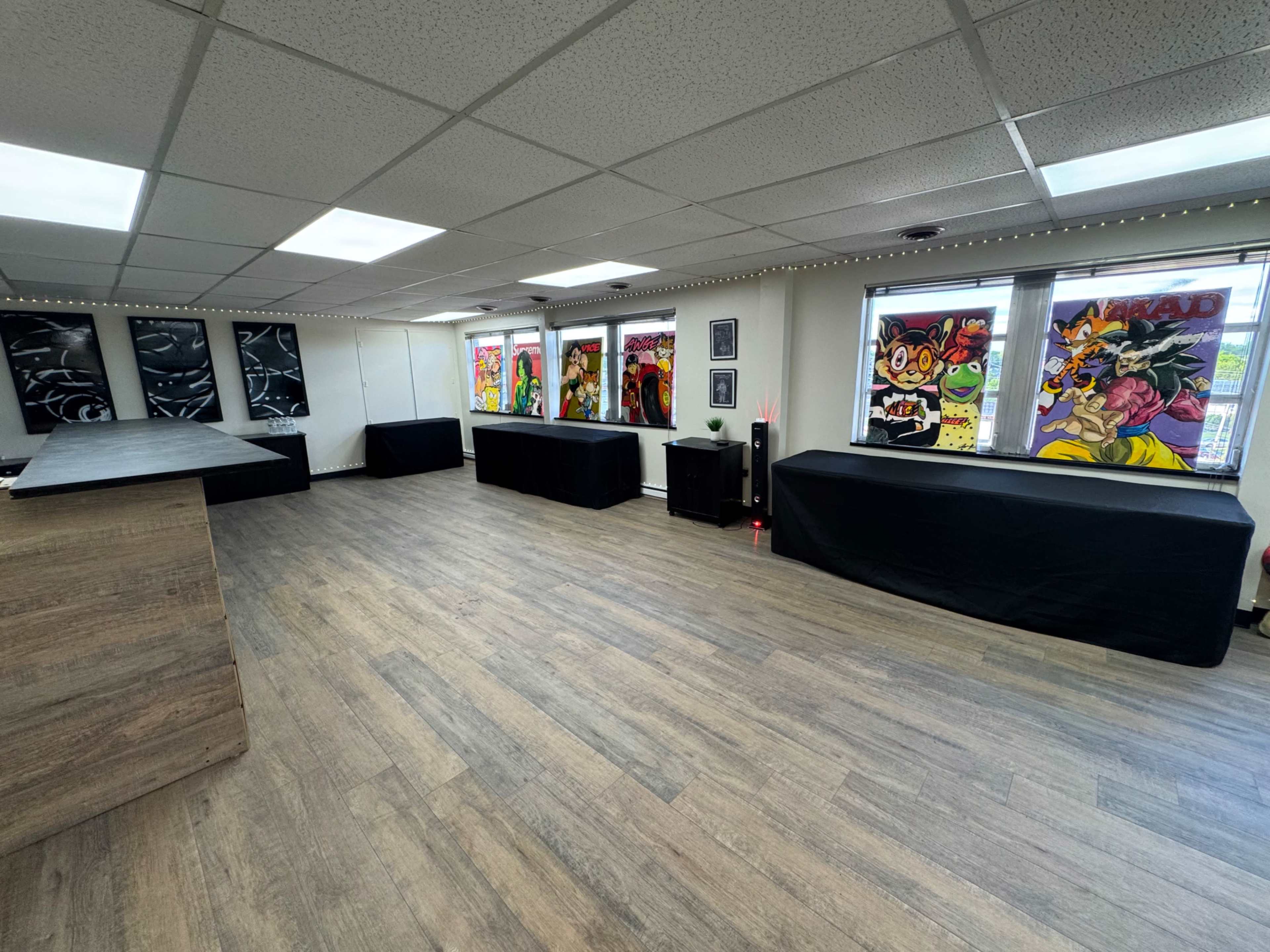 The image shows an empty room with wooden flooring, several covered tables, and colorful artwork displayed in large windows.