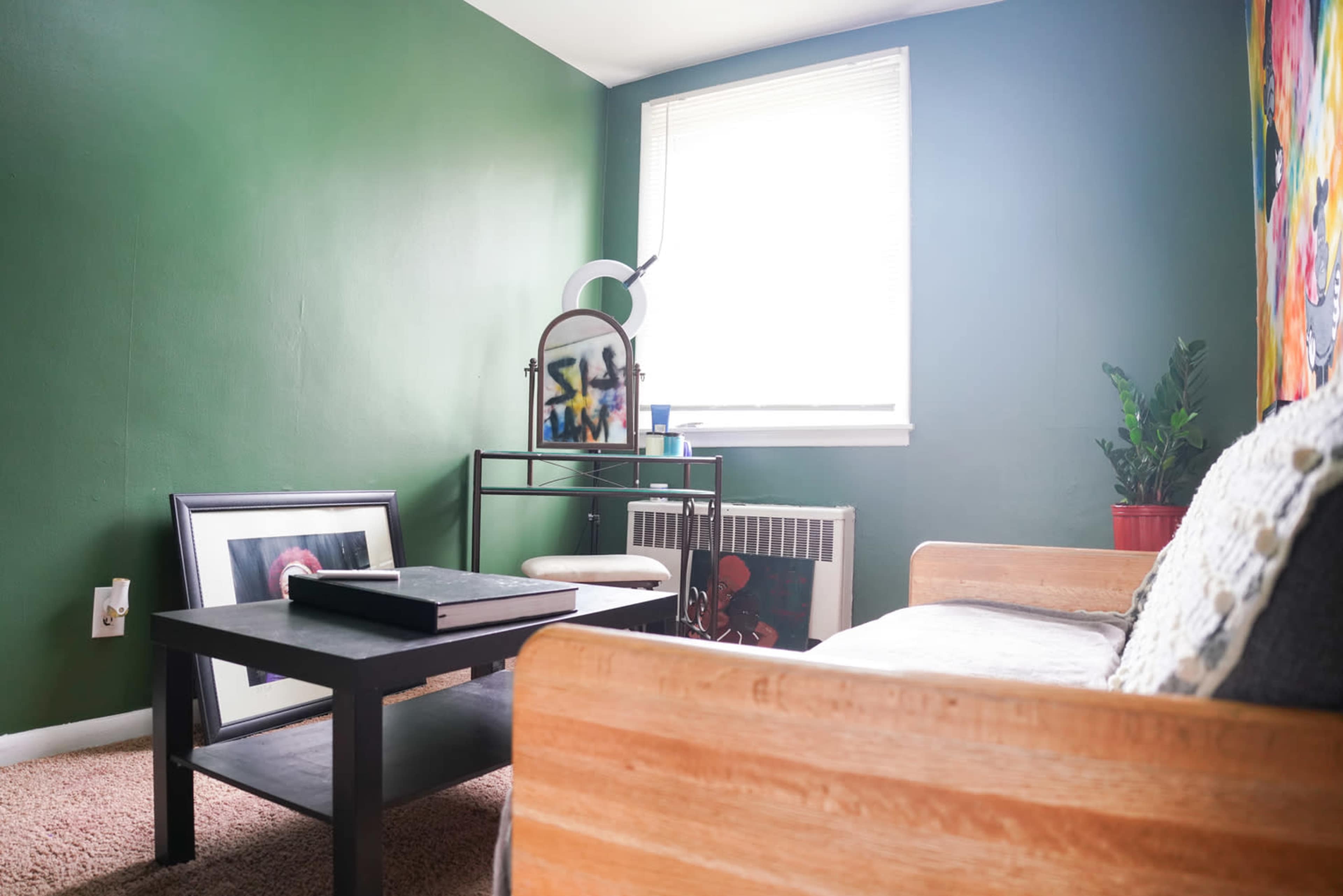 A room features a green wall, a wooden couch, a coffee table, an armchair, and a window with natural light.