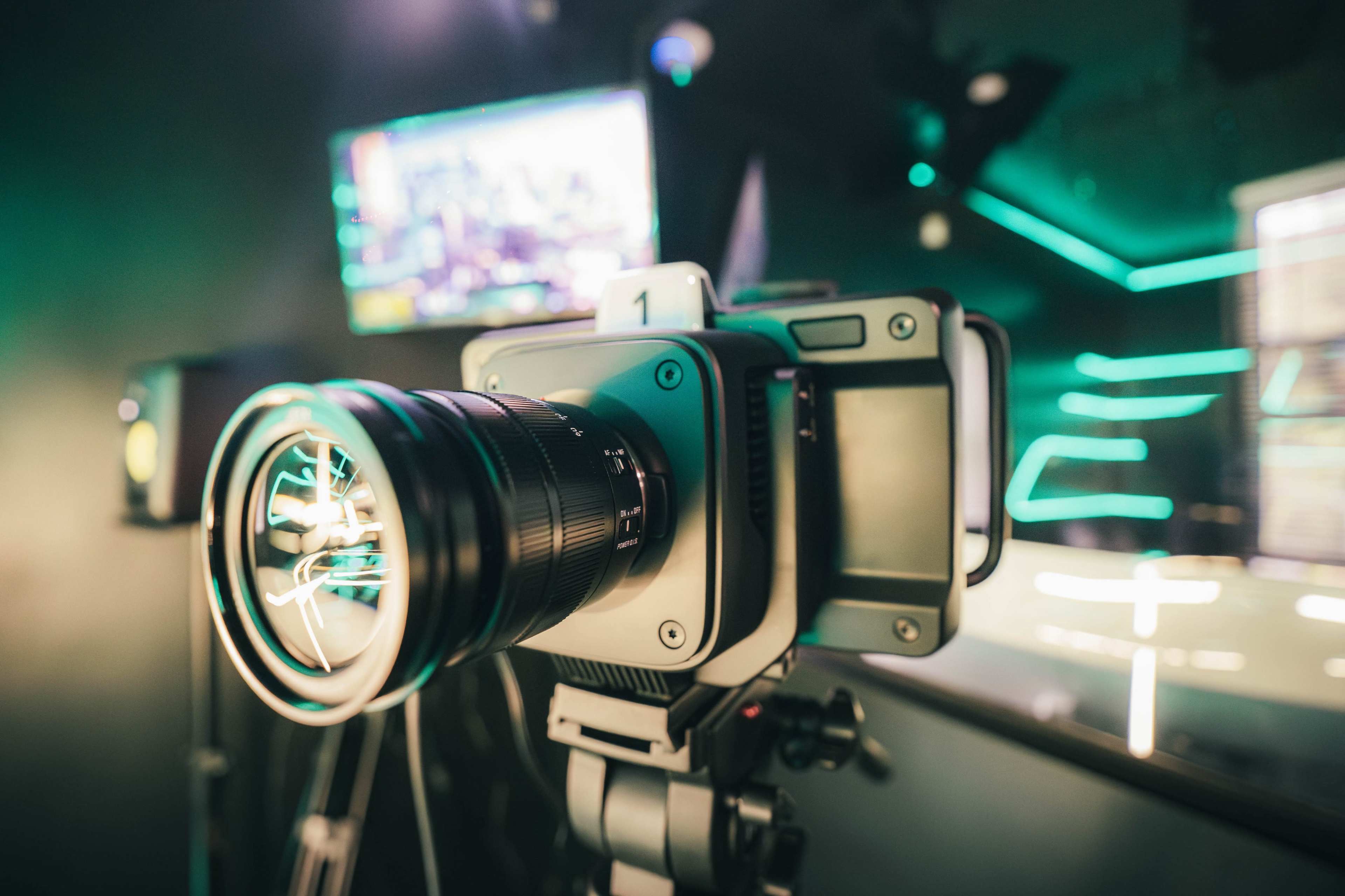 A professional camera is mounted on a tripod in a dimly lit room with green lighting and a screen in the background.