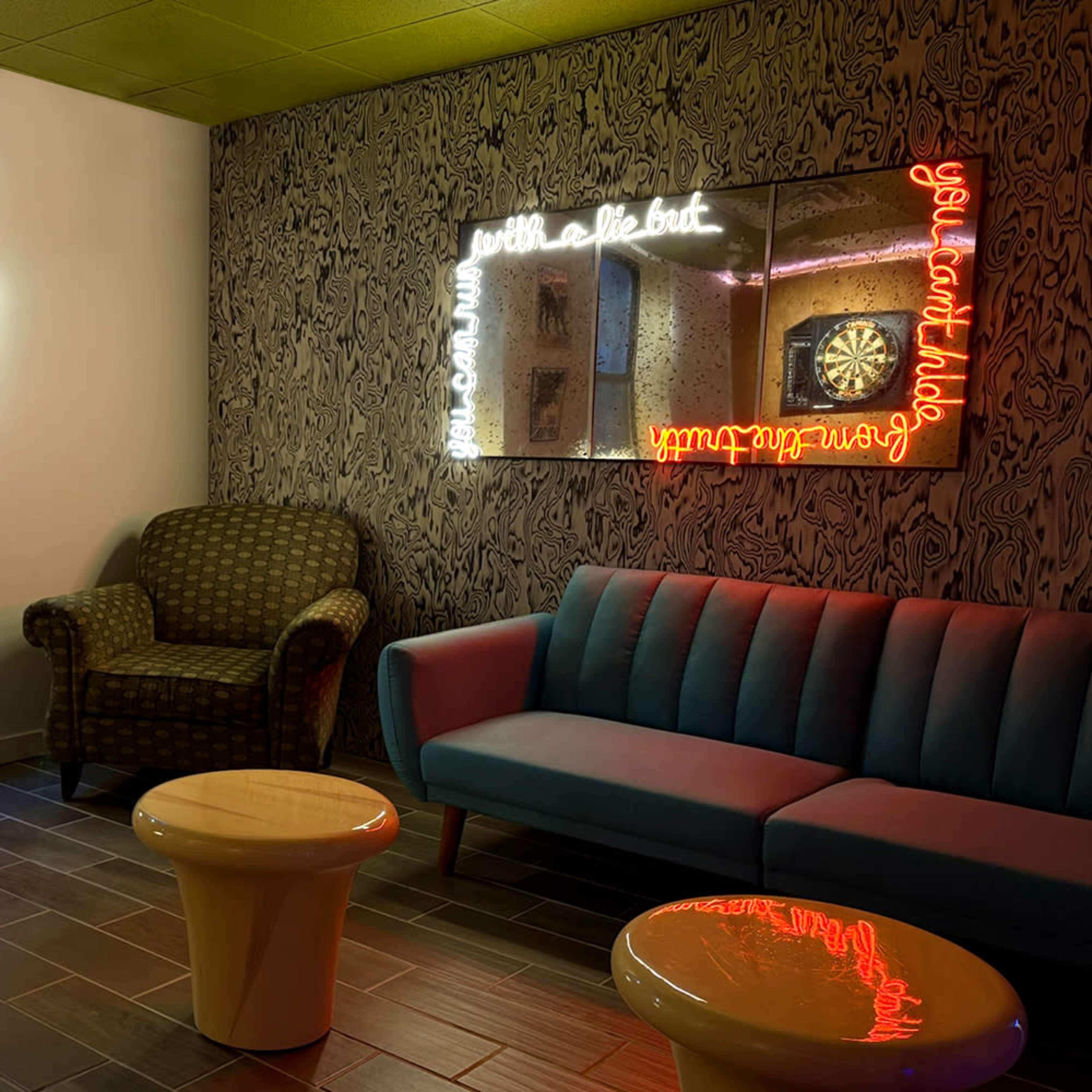 The image shows a cozy corner lounge with a patterned wall, featuring a green armchair, a blue sofa, and two white side tables, alongside a mirror illuminated by neon signs.