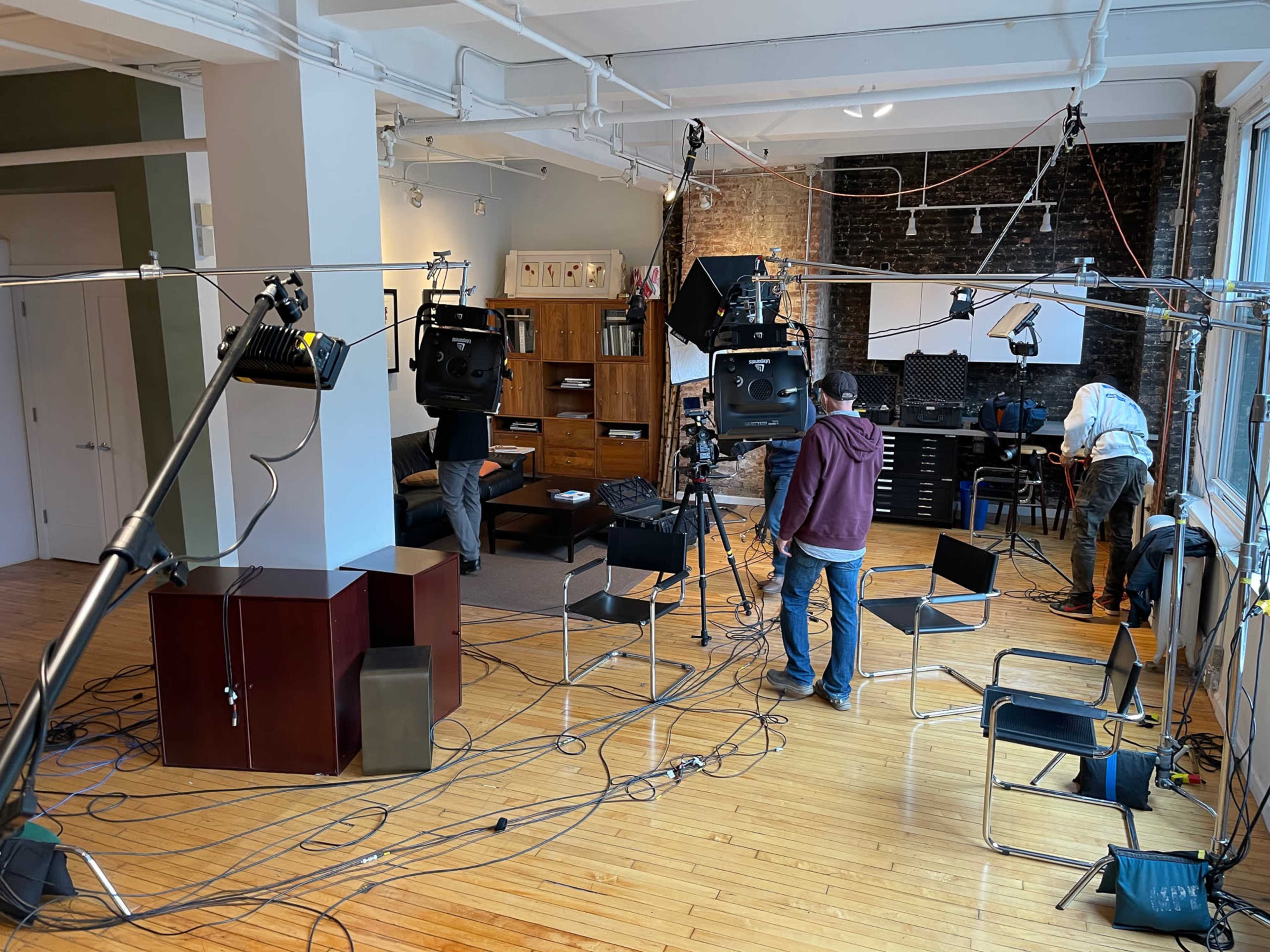 A production crew is setting up lighting and equipment in a spacious studio with wooden floors and a brick wall.