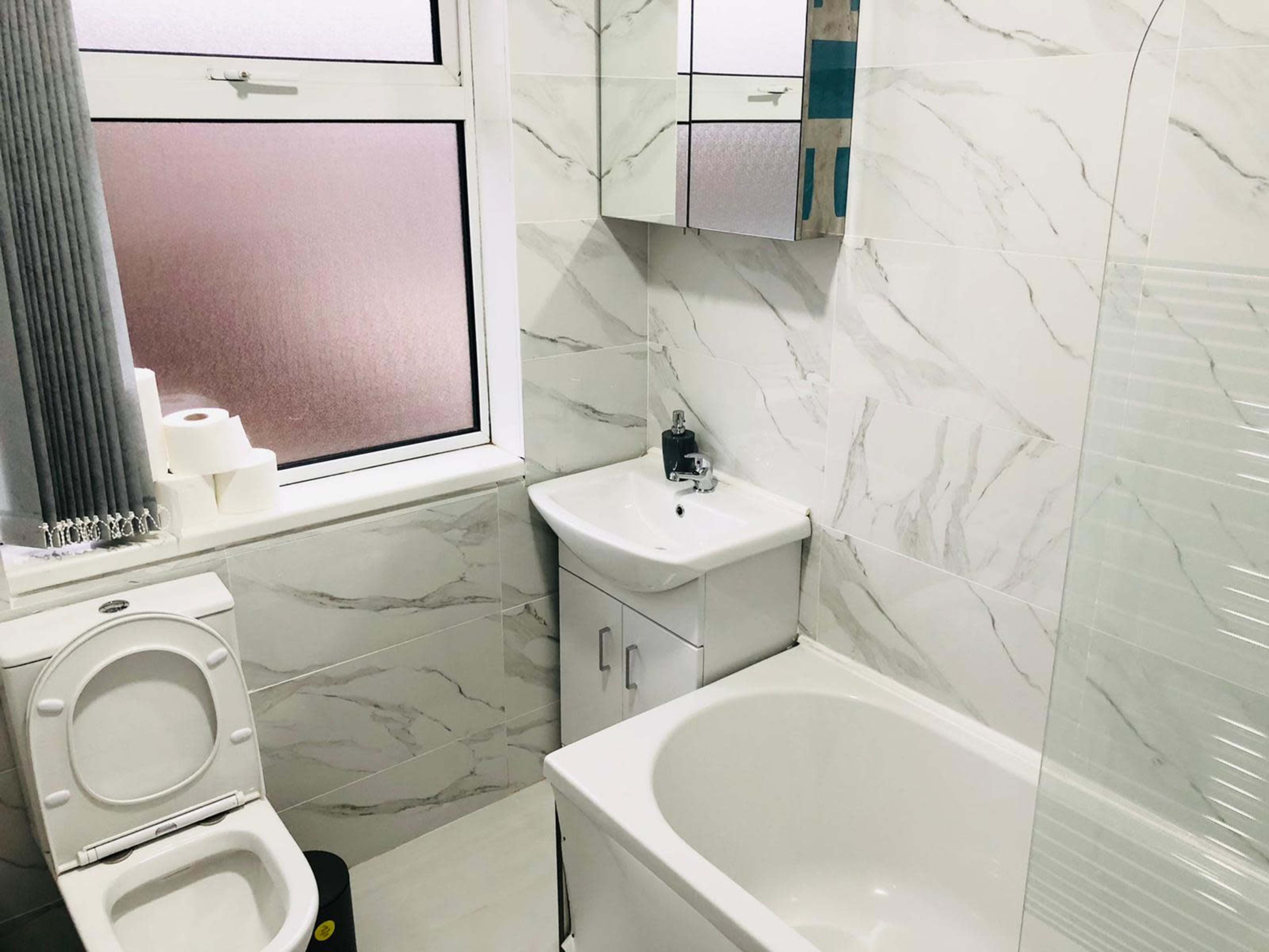 A modern bathroom features a white toilet, a basin with a mirror cabinet above, a bathtub, and a large window with sheer curtains.