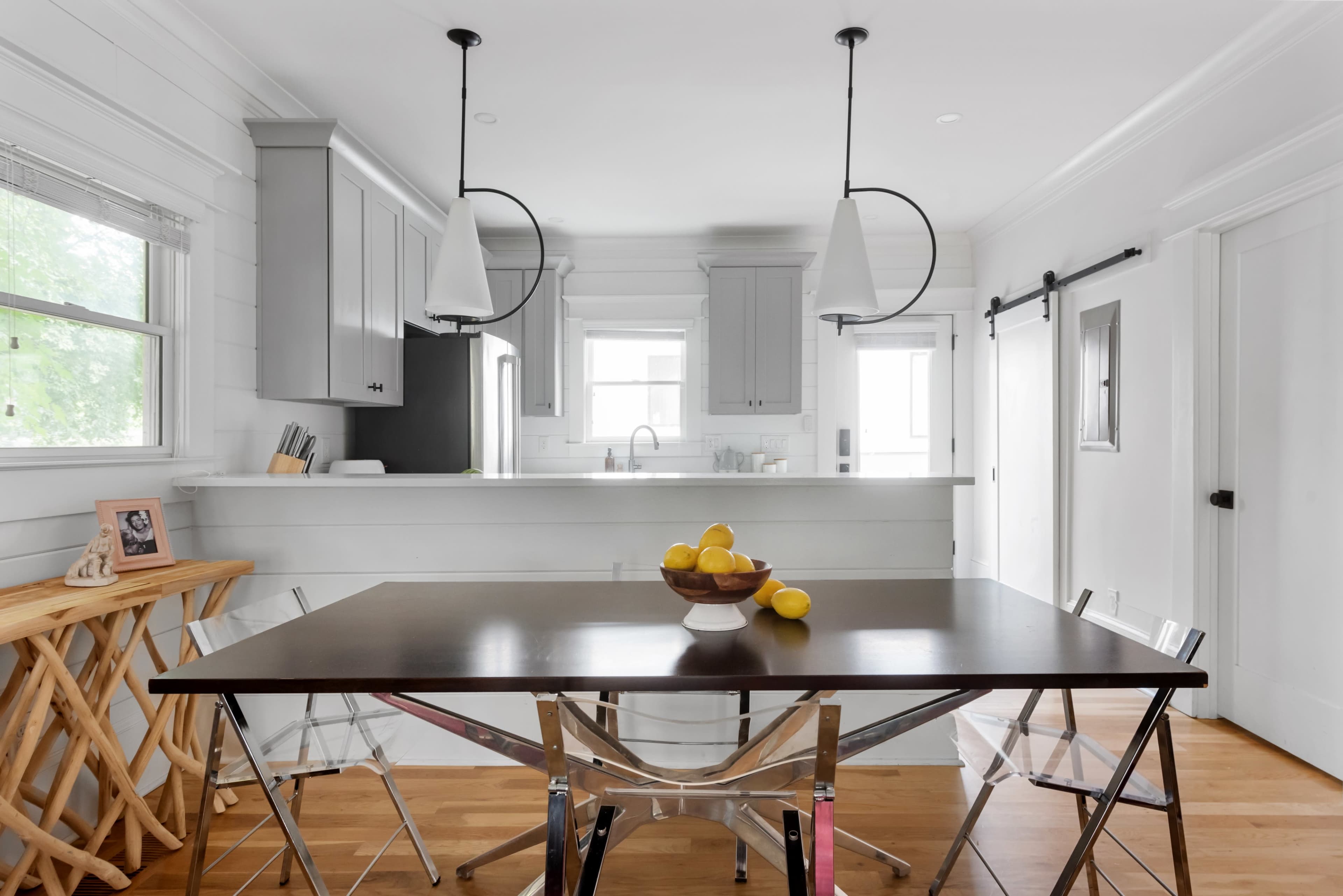 A modern dining area features a dark wooden table with a bowl of lemons at its center, surrounded by clear chairs, with a minimalist kitchen visible in the background.