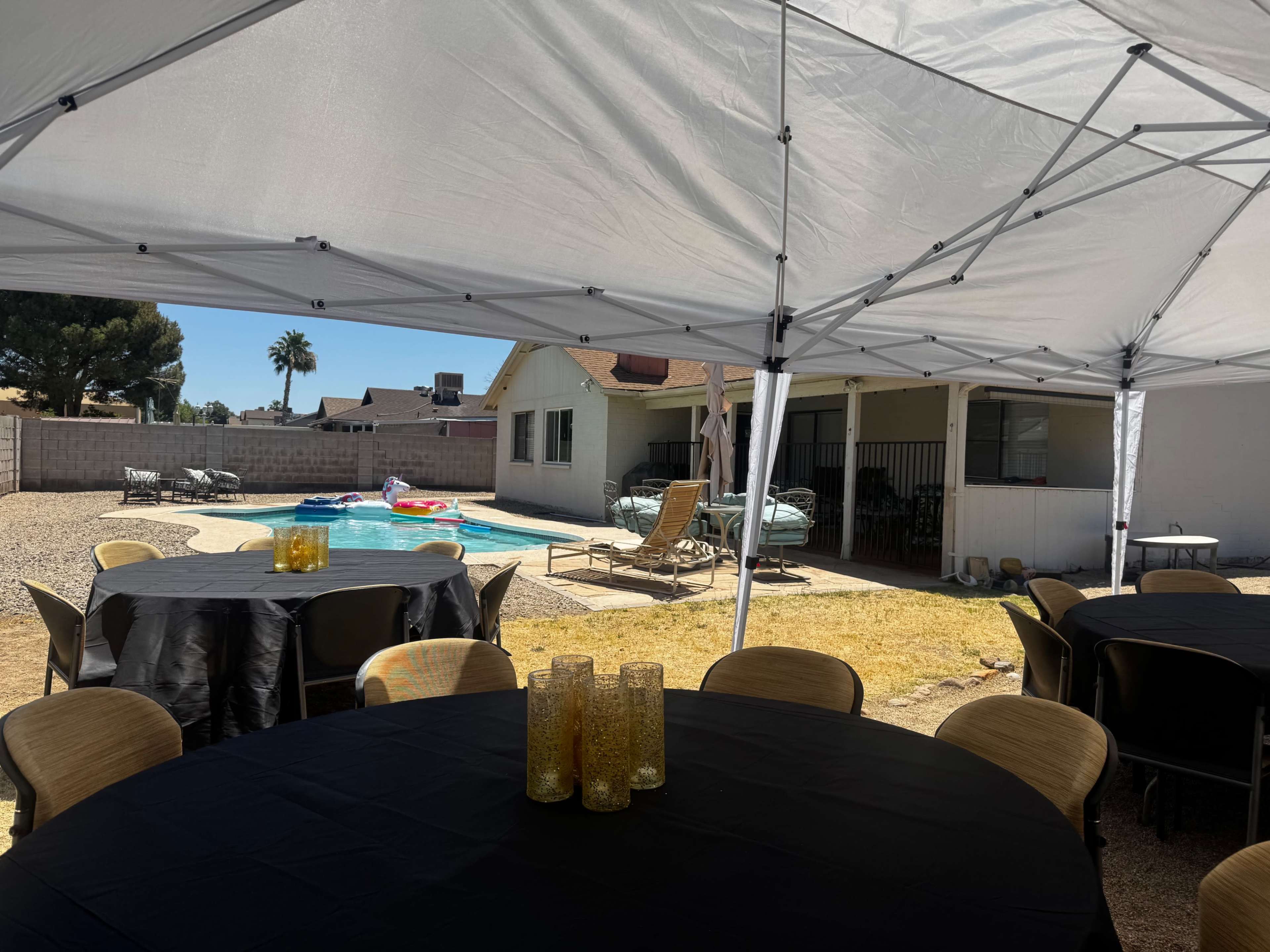 A clear tent shelters round tables near a swimming pool surrounded by lounge chairs and a house in the background.