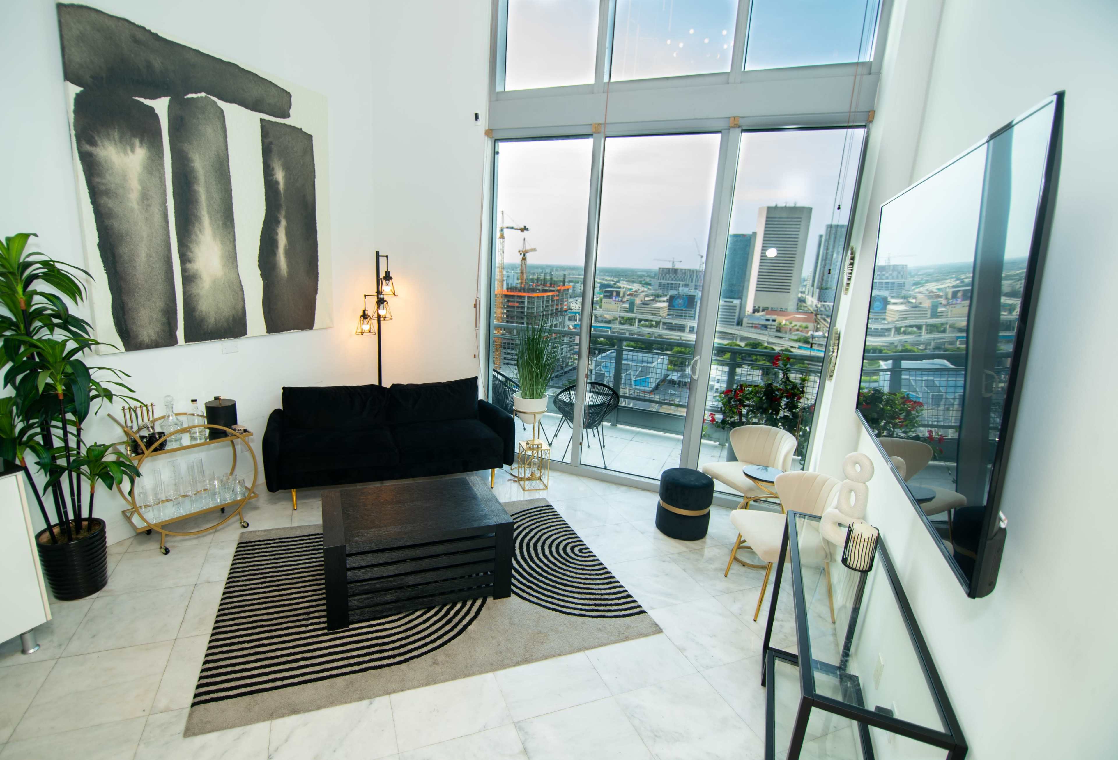 A modern living room with large windows offering a view of the city, featuring a black sofa, a geometric rug, a coffee table, and decorative plants.