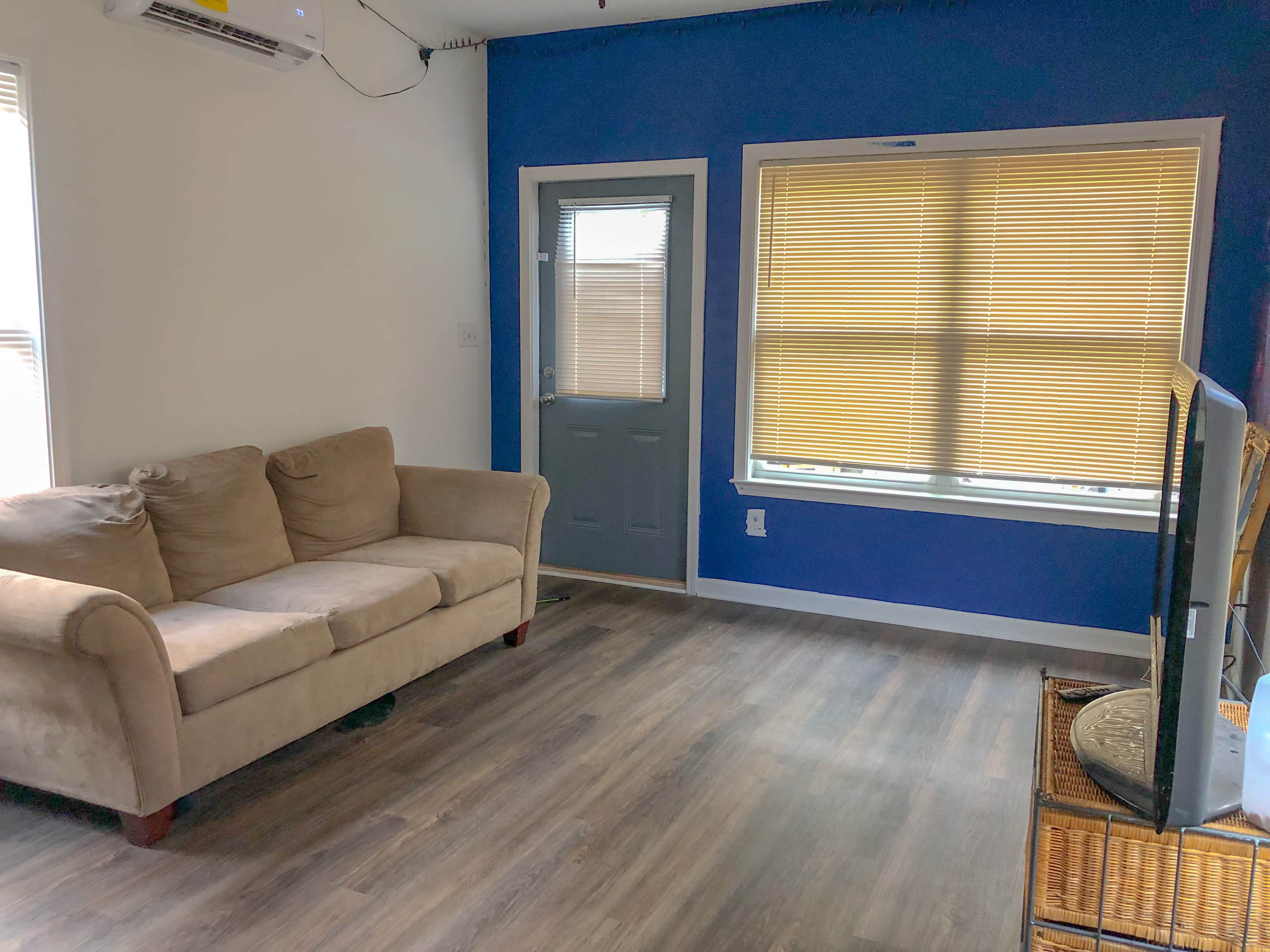 The image shows a living room with a beige sofa, a door with glass panels, a window with yellow blinds, and a television on a stand.