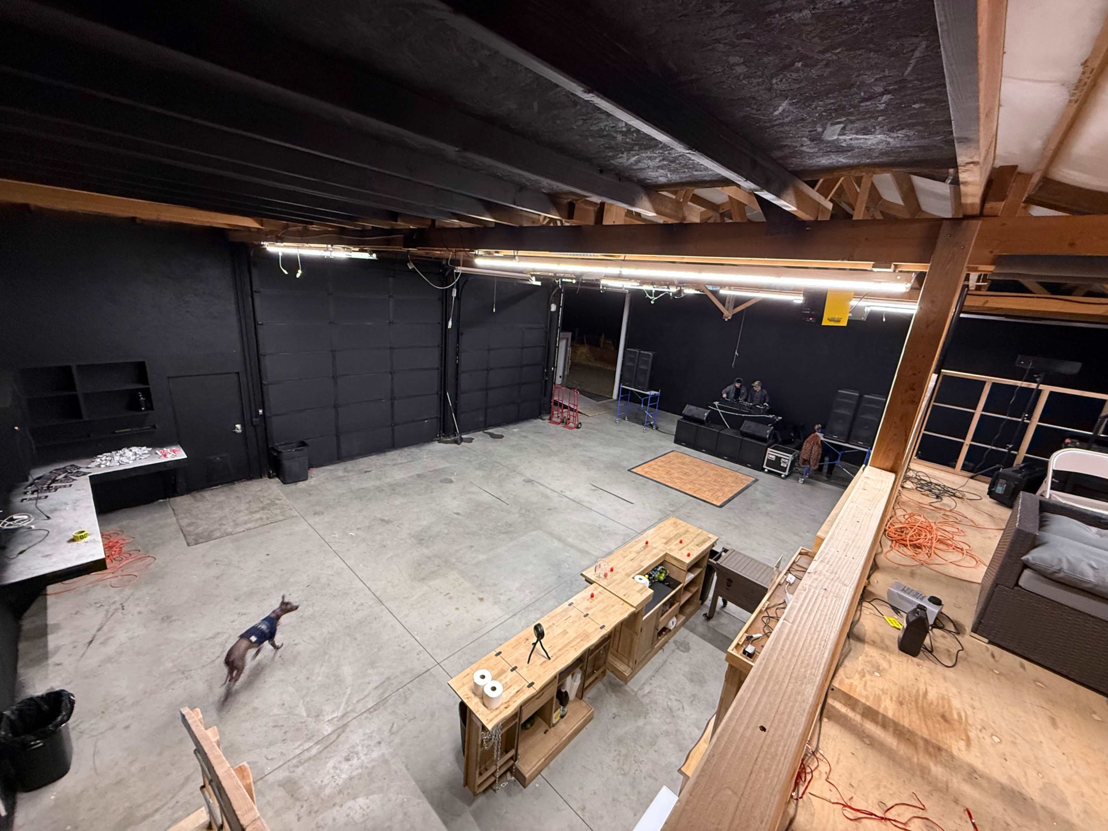 The image shows an empty indoor workspace with wooden tables, a concrete floor, and two people in the background, while a dog walks across the space.