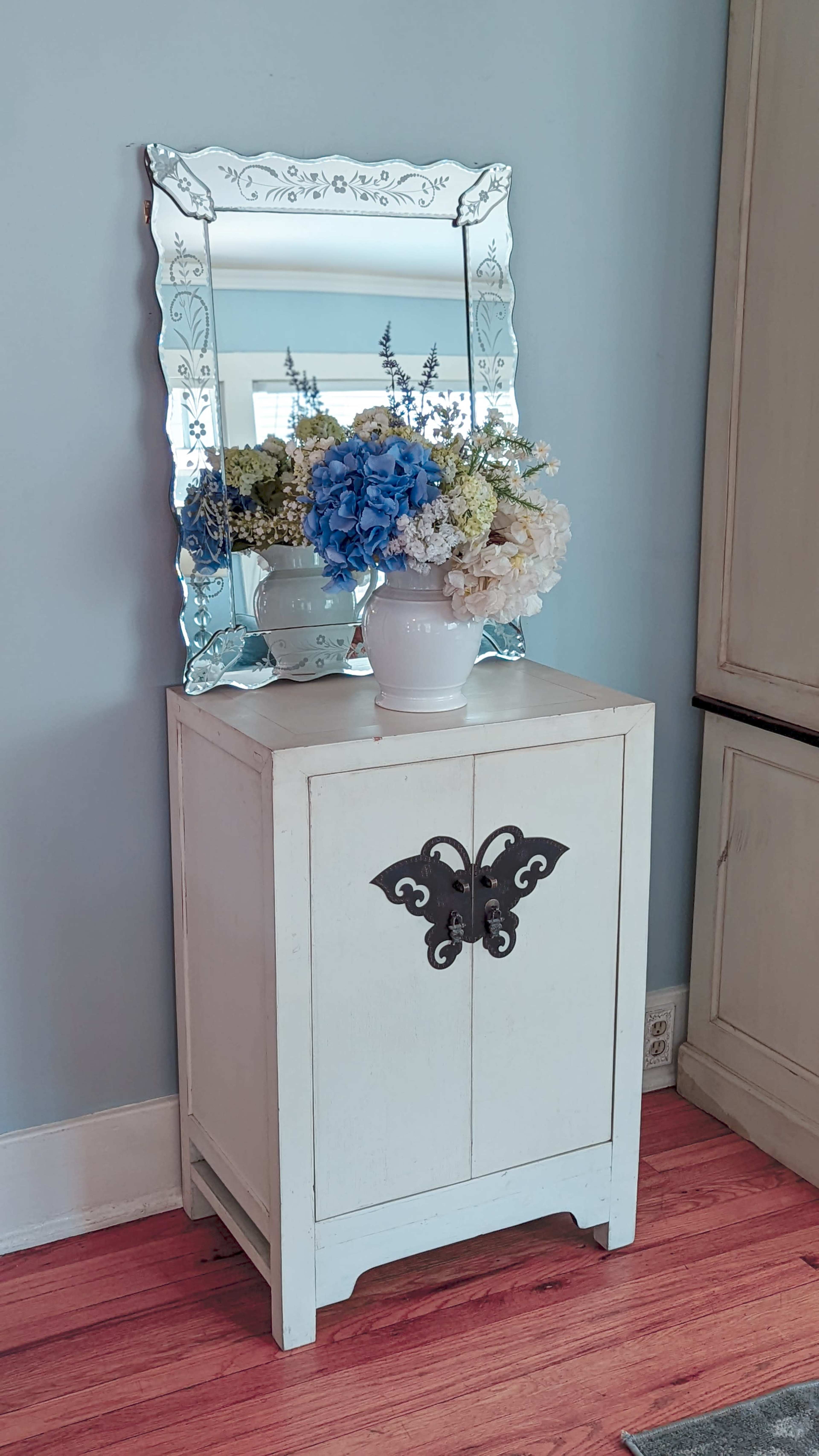 A white cabinet with a butterfly design in front of a mirror displays a vase of blue and white flowers.