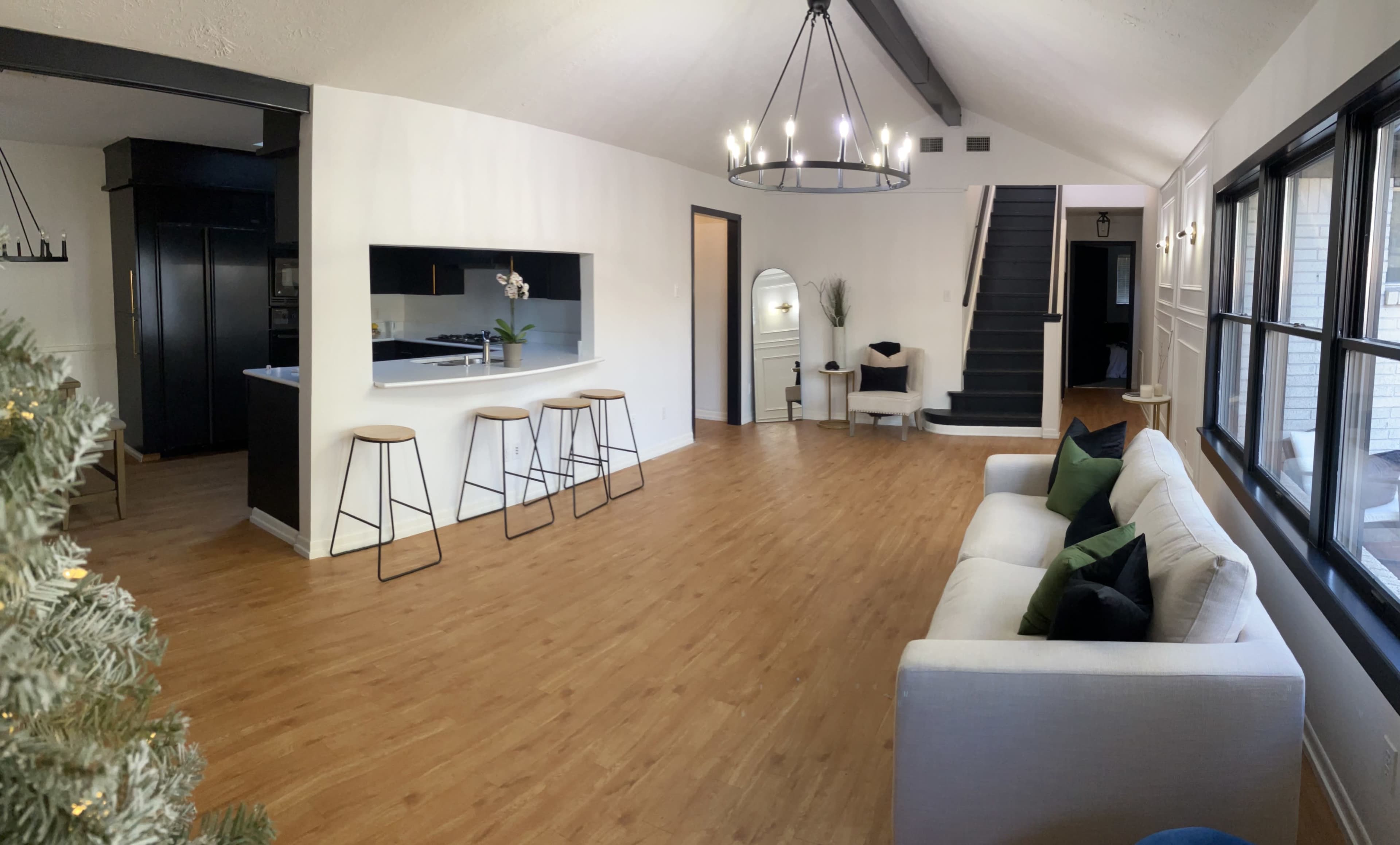 A spacious, well-lit living area featuring a white sofa, wooden flooring, bar stools at a counter, and a staircase leading to another level.