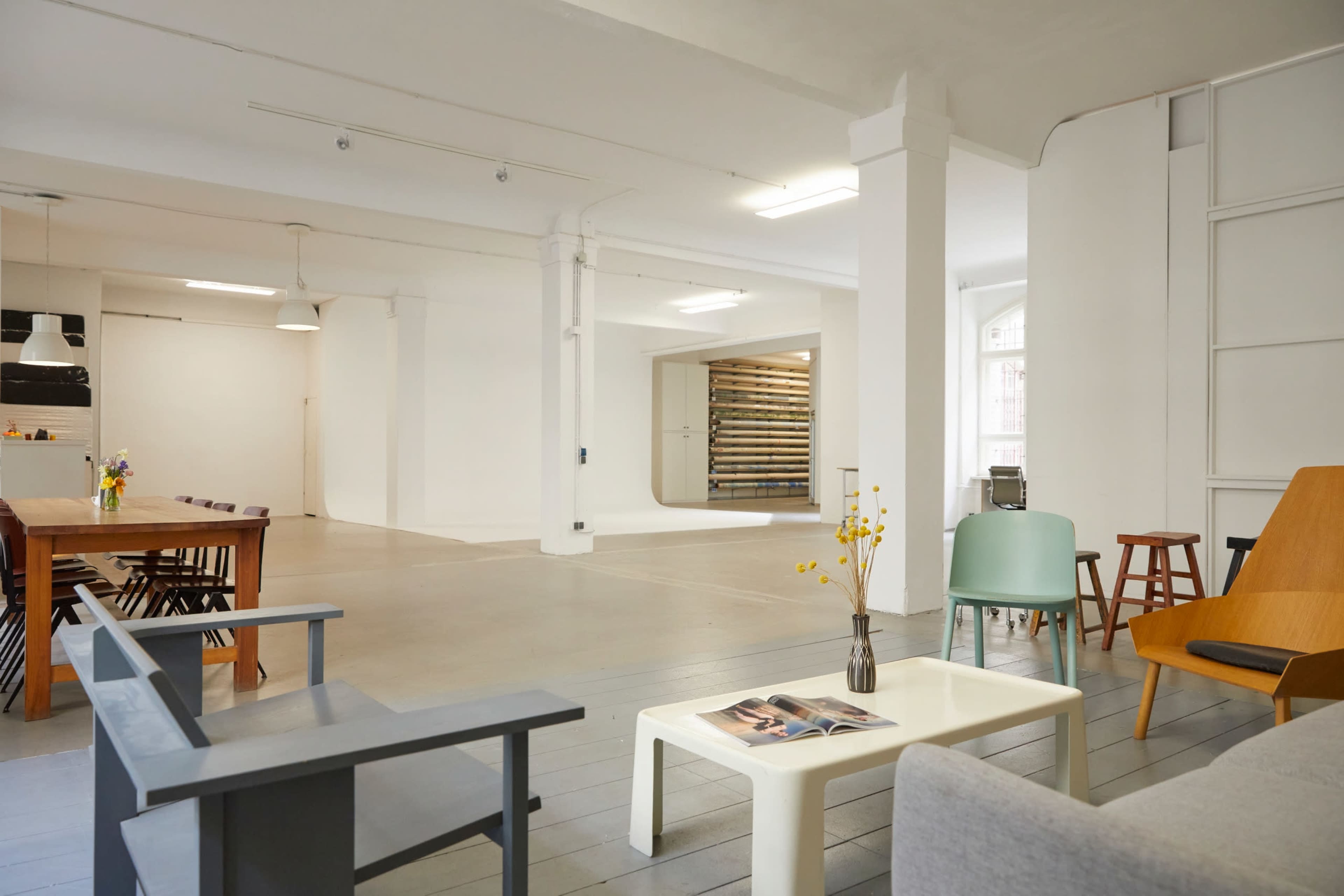 A spacious, brightly lit room with a minimalist design featuring various seating arrangements, a dining table, and a wooden storage area in the background.