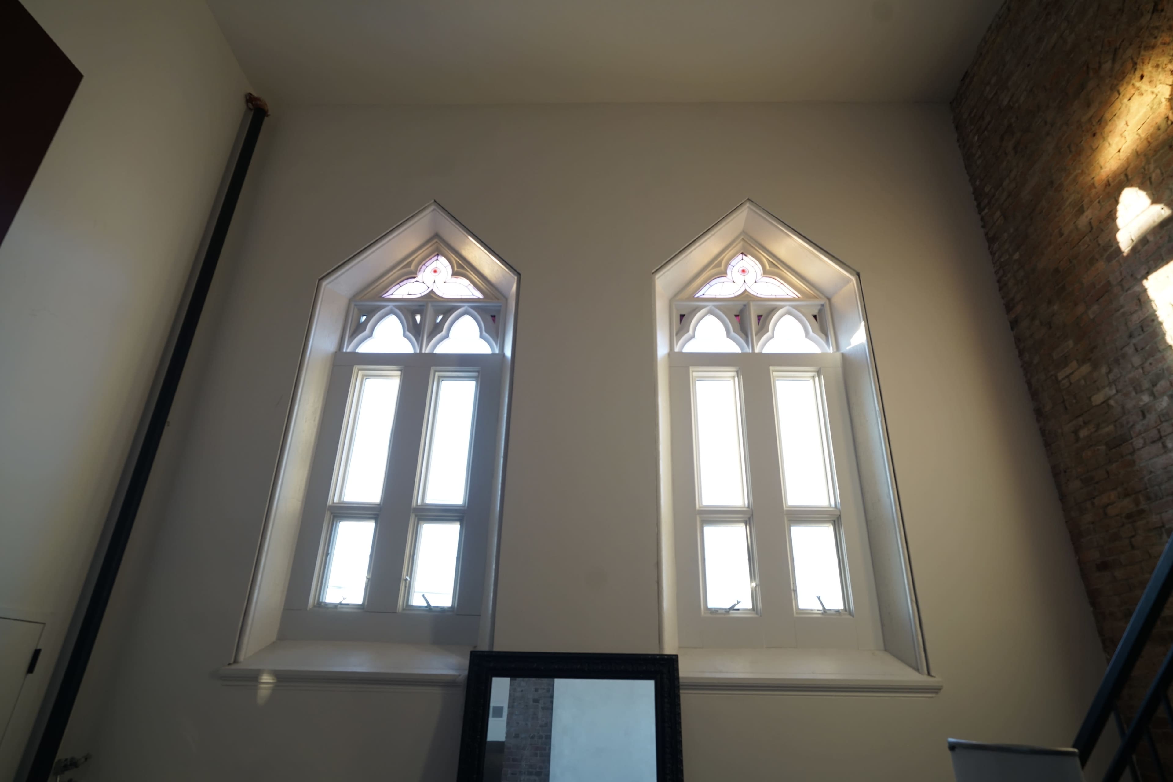 Two Gothic-style windows with pointed arches and decorative elements, set in a light-colored wall, accompanied by a large mirror on the floor below.