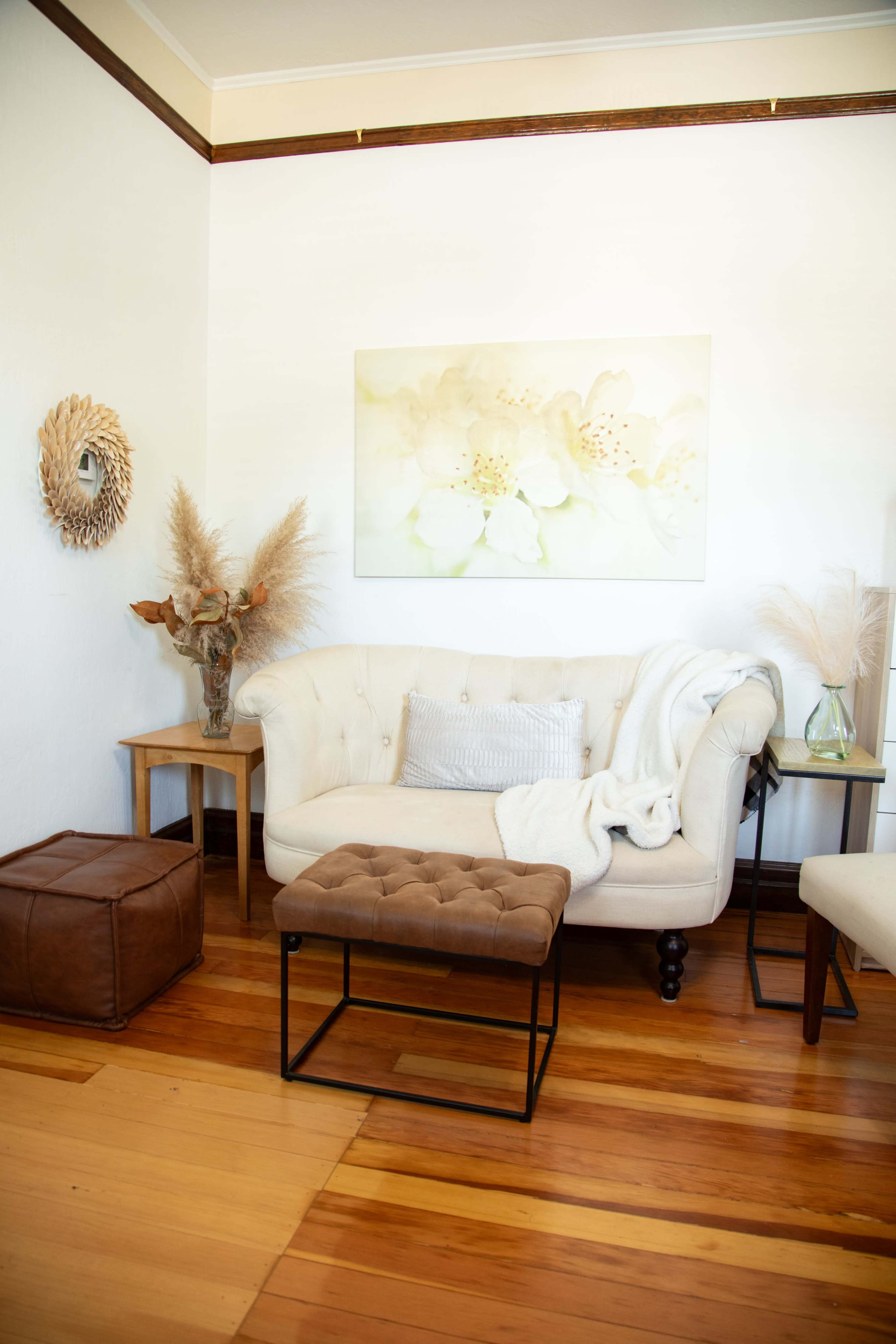 A cozy living room features a tufted beige sofa, a decorative wall hanging, a small side table with a vase of dried flowers, and a leather pouf on wooden floors.