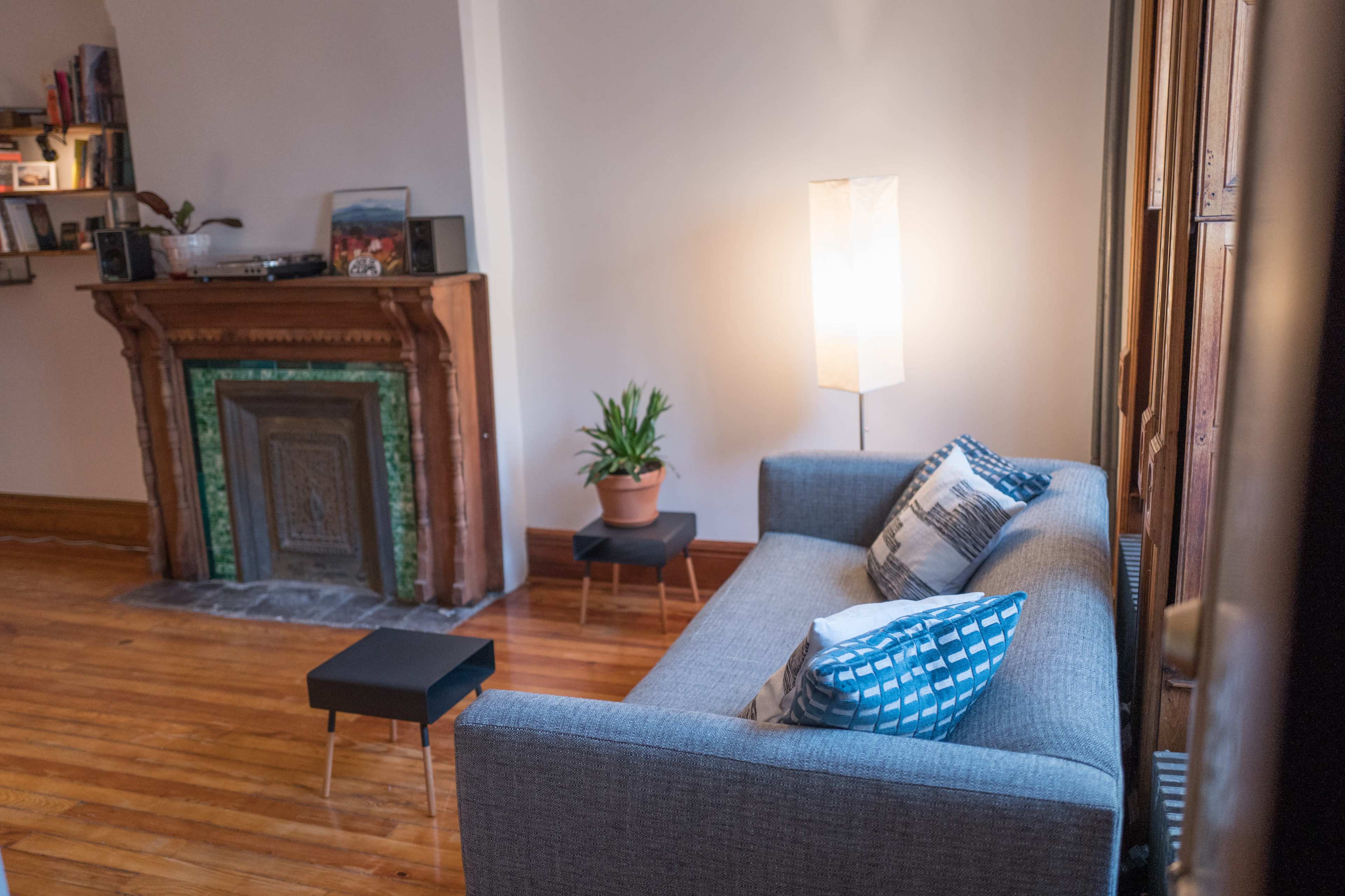 A gray sofa with patterned cushions sits across from a wooden fireplace, accompanied by a potted plant and a tall lamp in a well-lit room with hardwood floors.