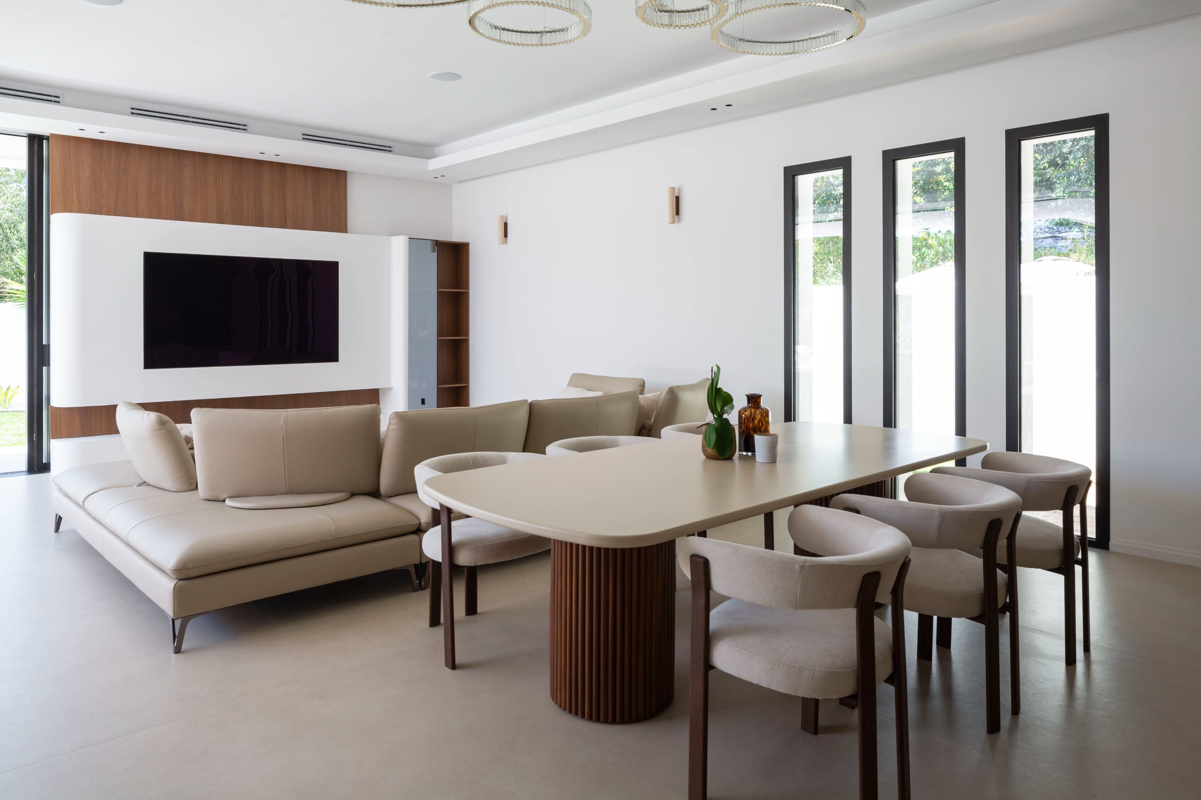 A modern living and dining area features a beige sectional sofa, a round dining table surrounded by upholstered chairs, and large windows allowing natural light to fill the space.
