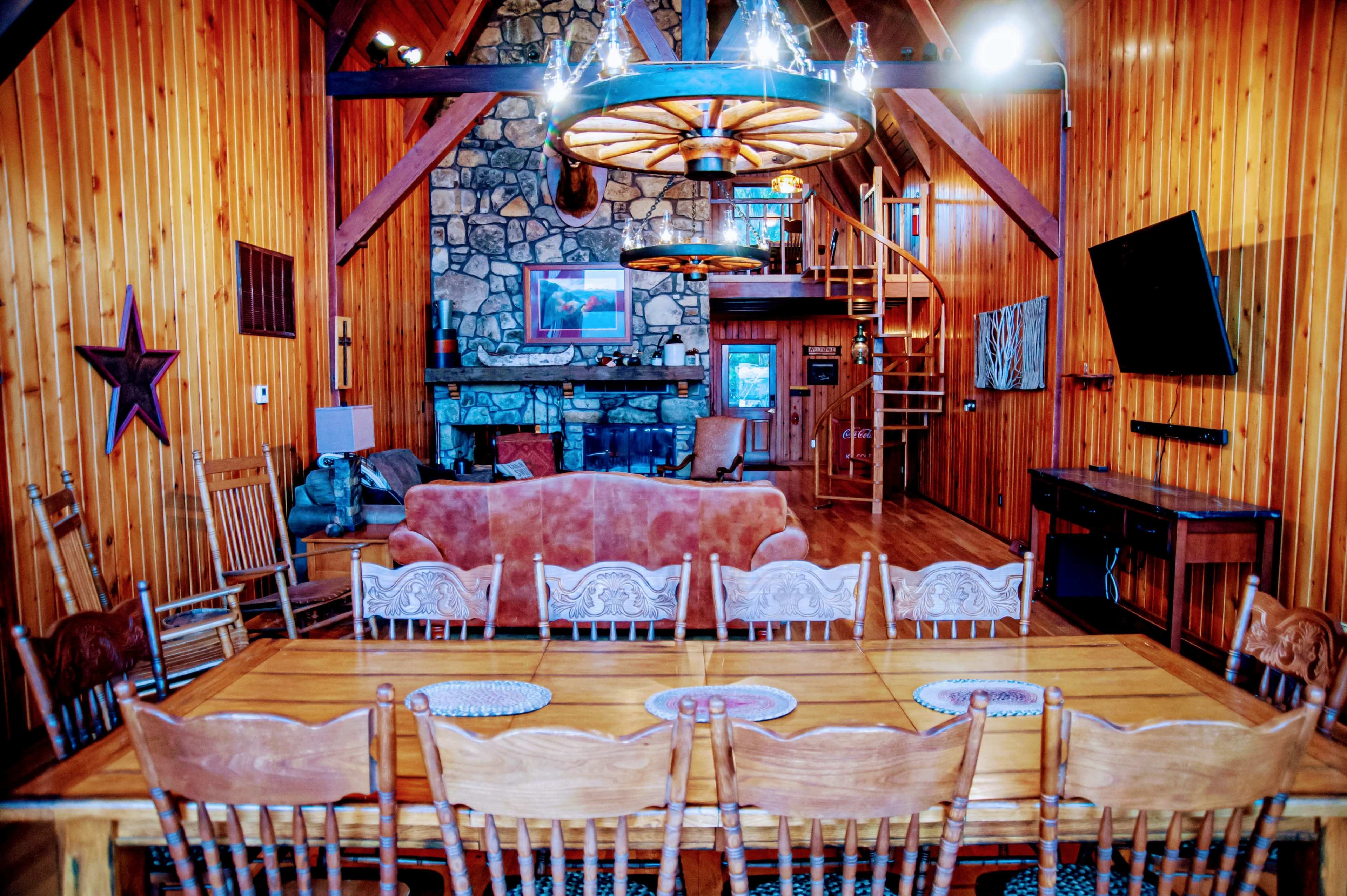 A wooden dining table surrounded by chairs is set in a rustic cabin living area featuring a stone fireplace and a spiral staircase.