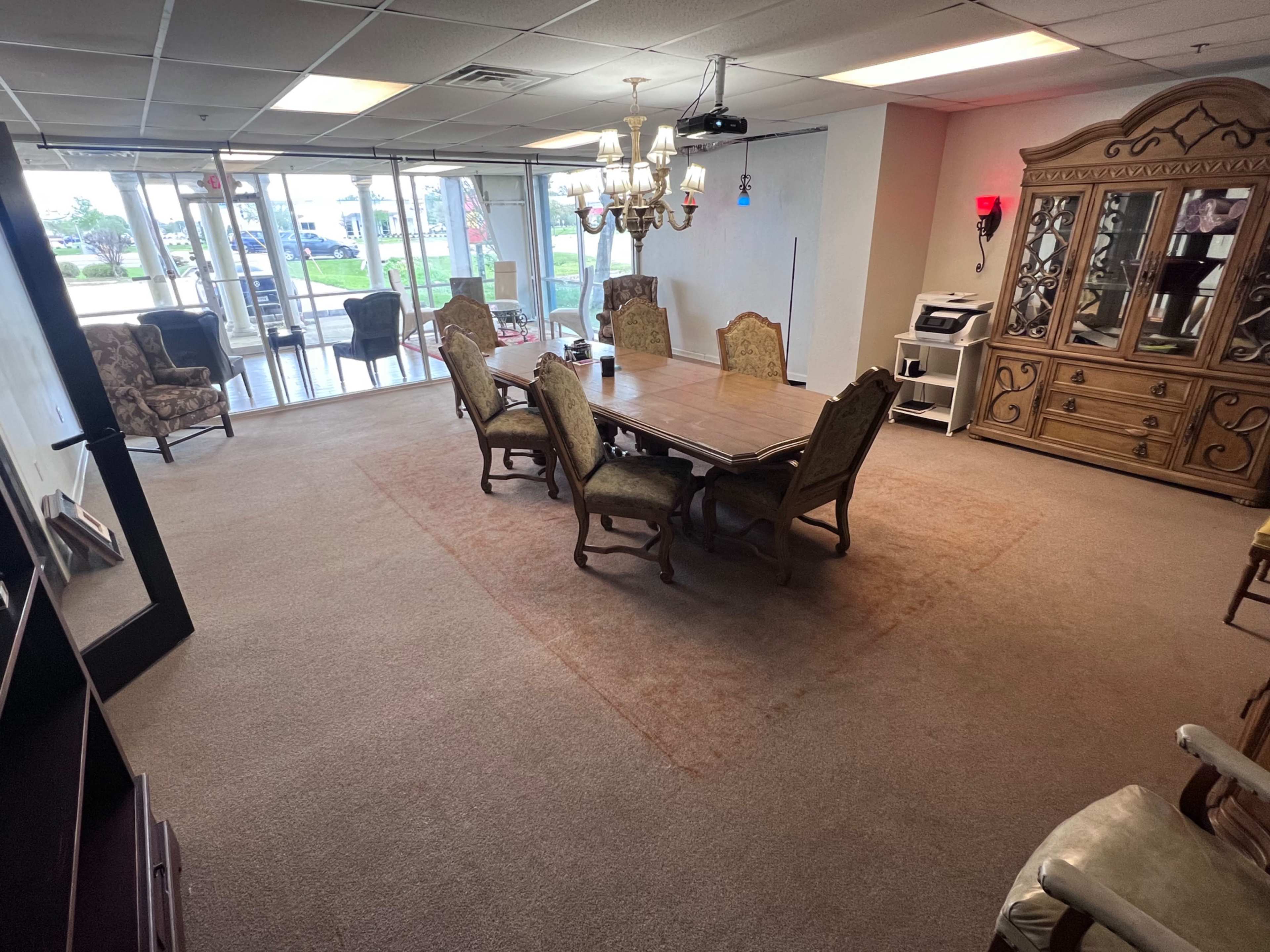The image shows a spacious, carpeted room with a large dining table surrounded by chairs, a chandelier overhead, and a cabinet against the wall.