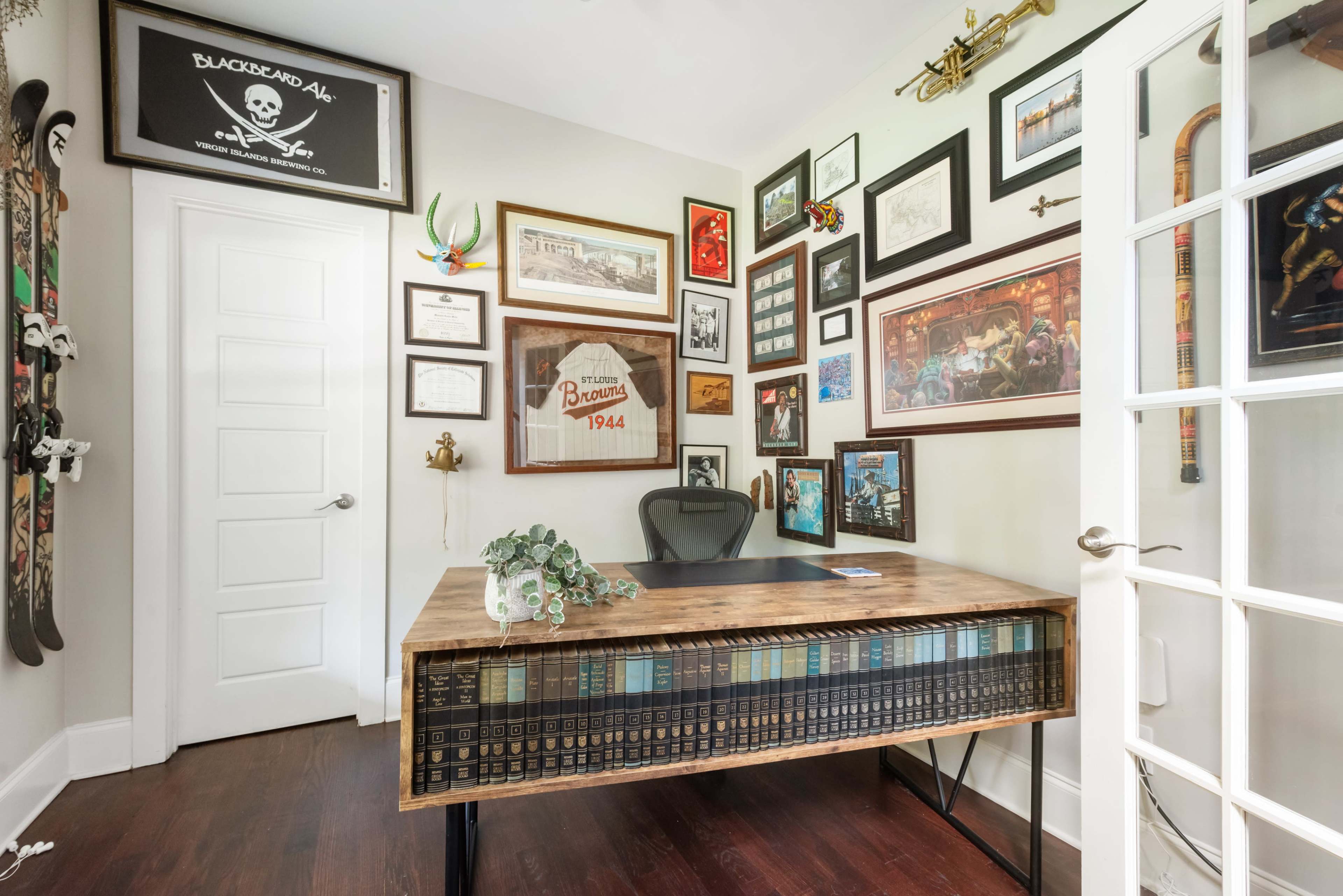 The image shows a home office with a wooden desk in front of a wall adorned with various framed pictures, certificates, and decorative items.