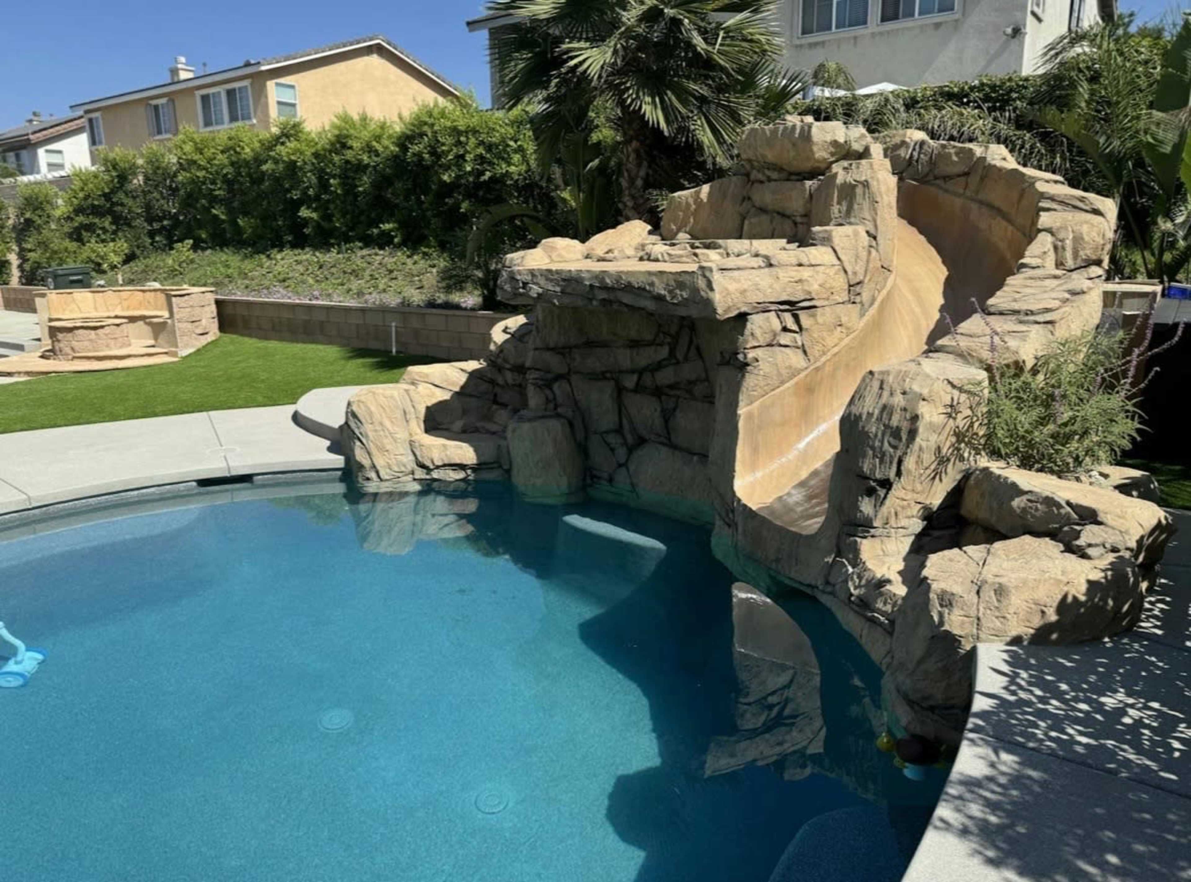 The image shows a swimming pool with a rock formation slide and tropical plants in the background.