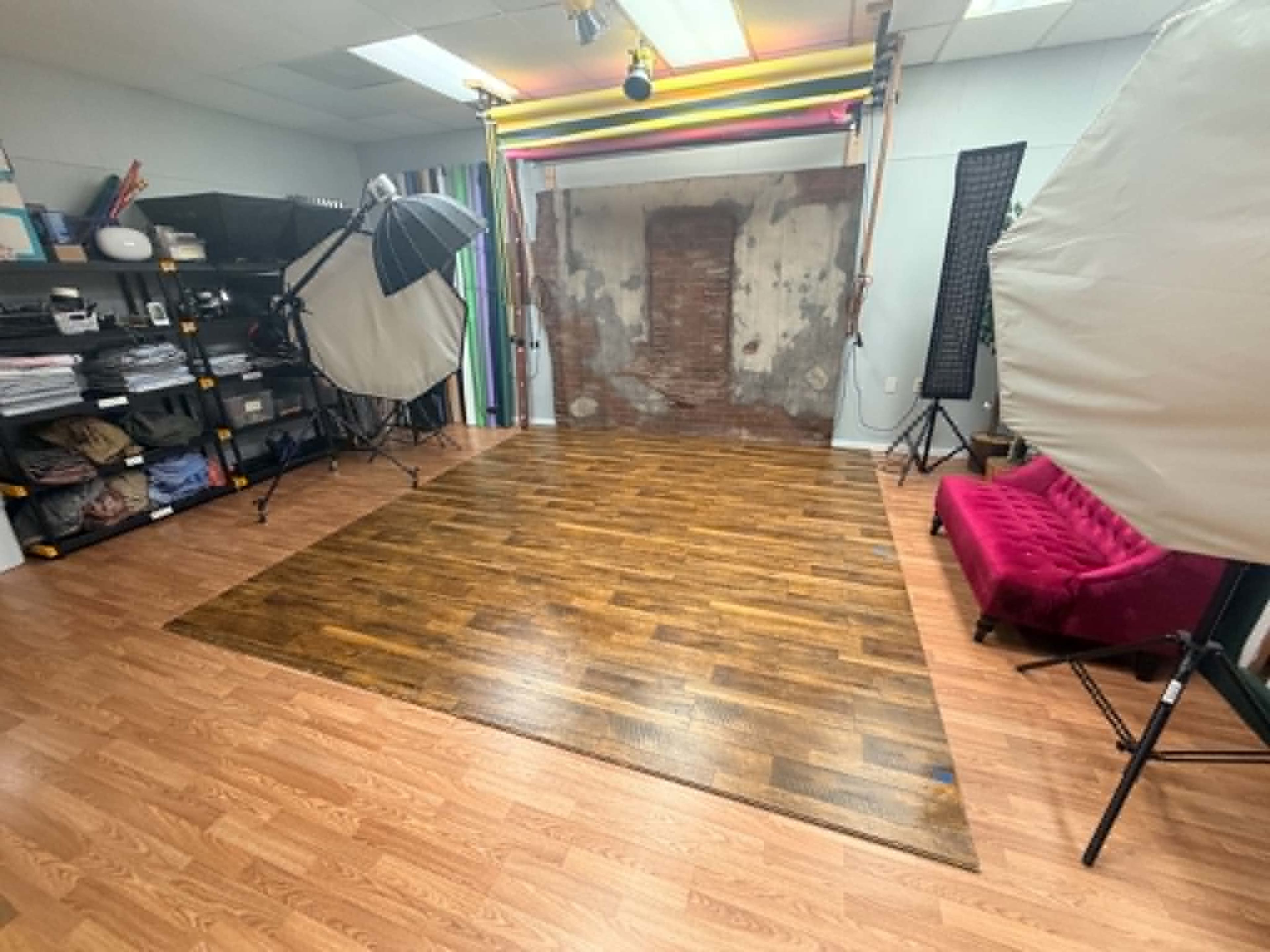 The image shows a photography studio with a hardwood floor, various lighting equipment, a colorful backdrop, and a bright red velvet couch.
