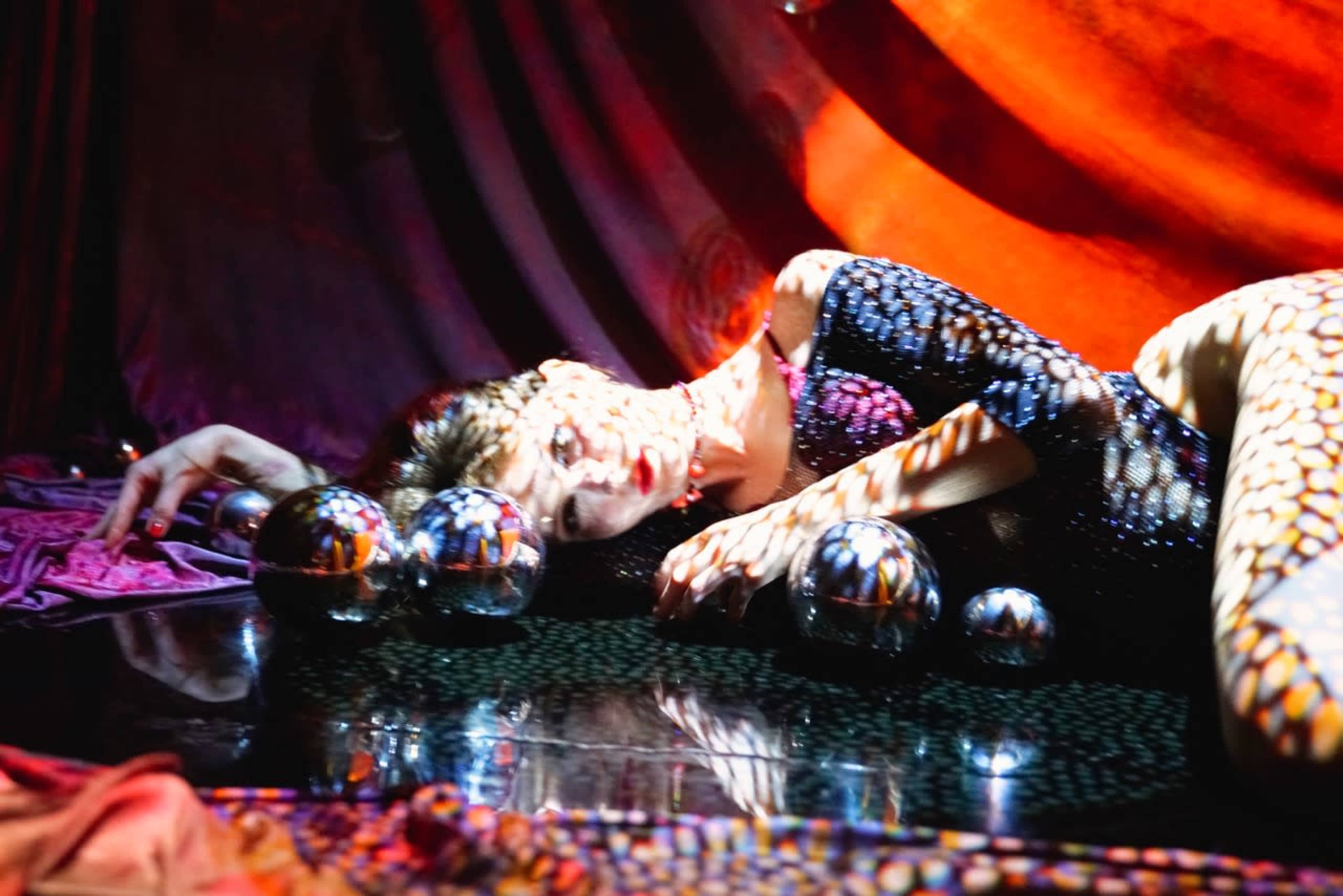 A person lies on a reflective surface surrounded by metallic spheres, with colorful light patterns playing across their body.