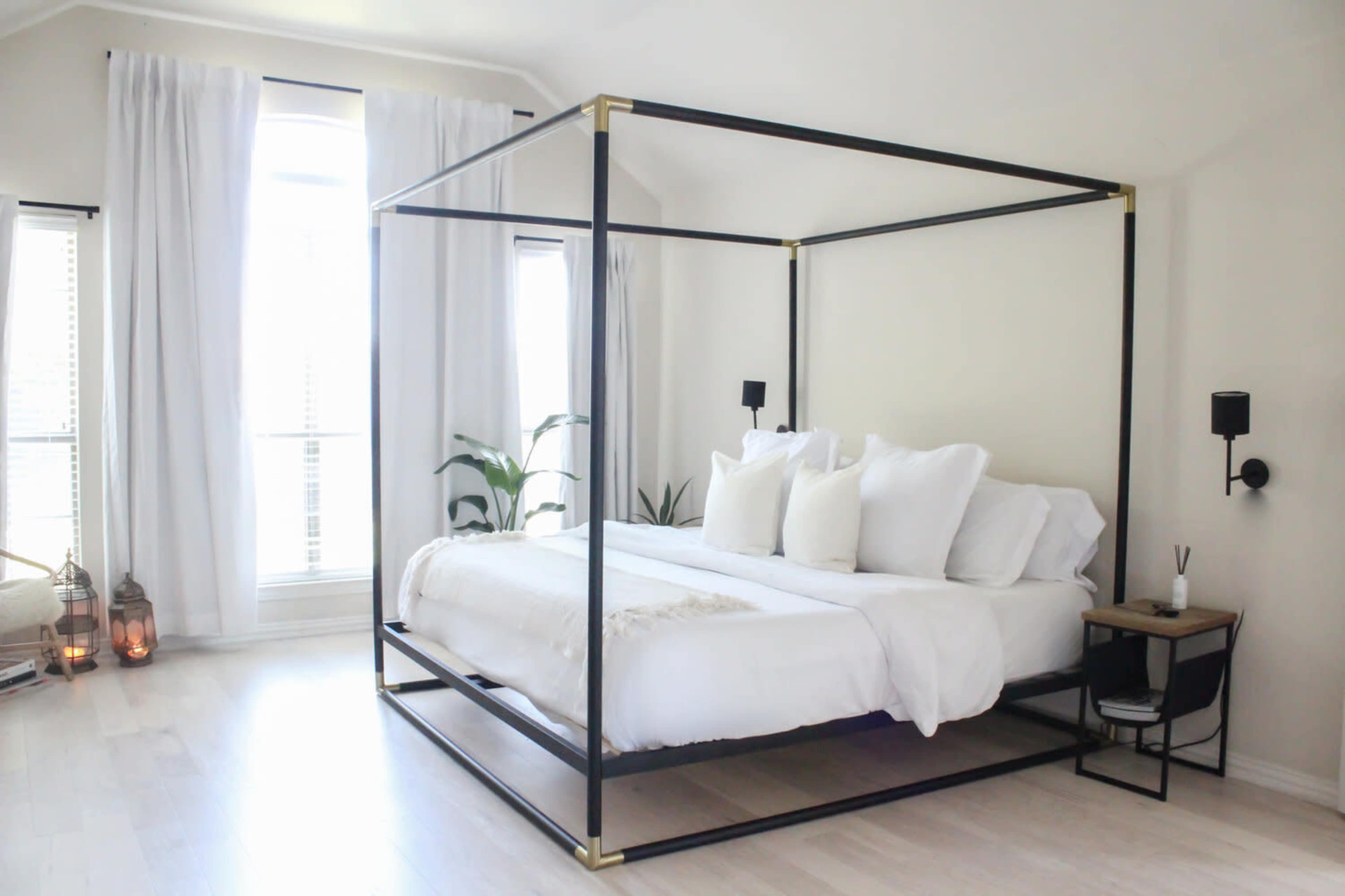 A modern bedroom featuring a black metal canopy bed with white bedding, flanked by large windows and decorative plants.