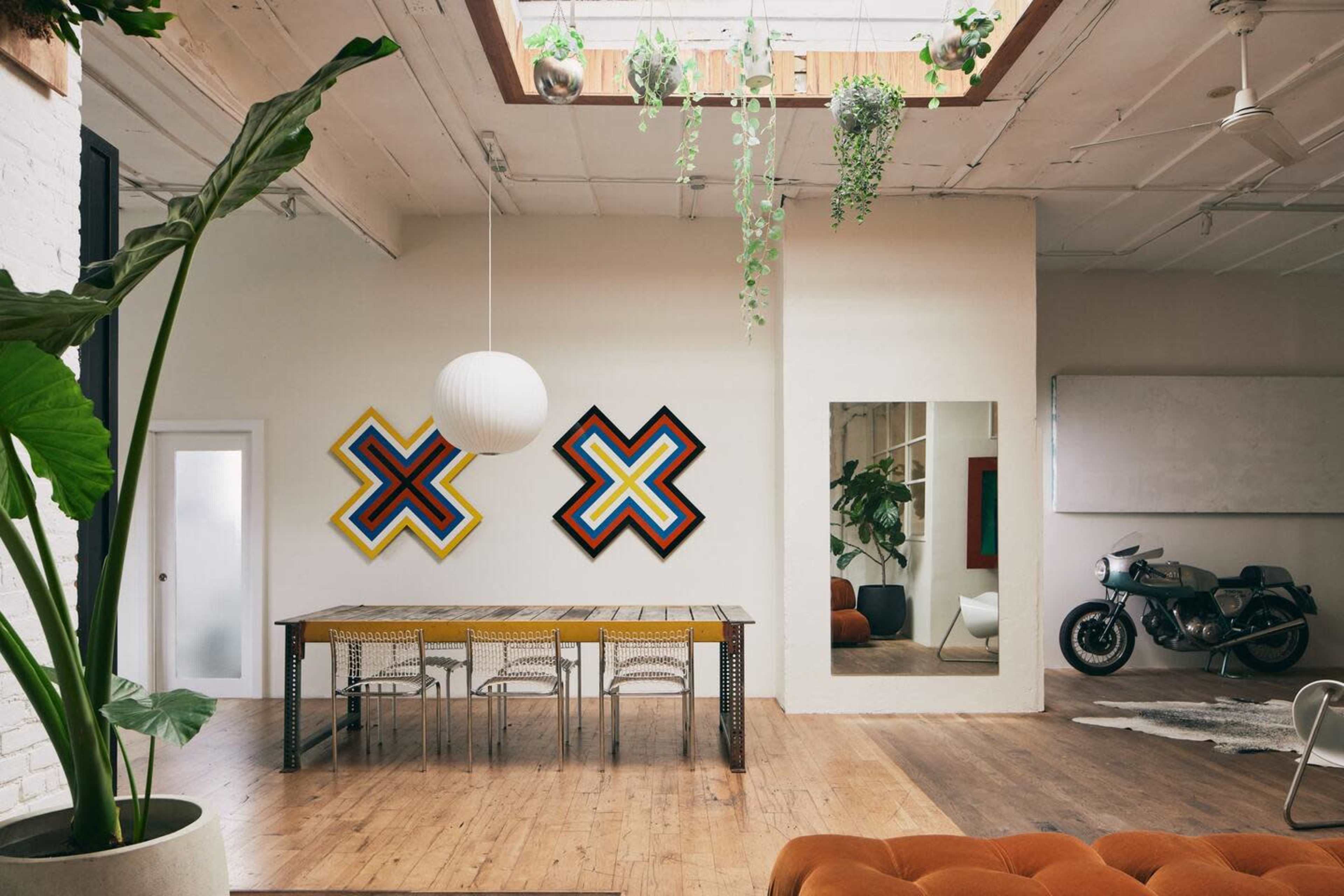 The image features a spacious room with a wooden dining table surrounded by chairs, two colorful geometric artworks on the walls, a large mirror, and a motorcycle beside a whiteboard.