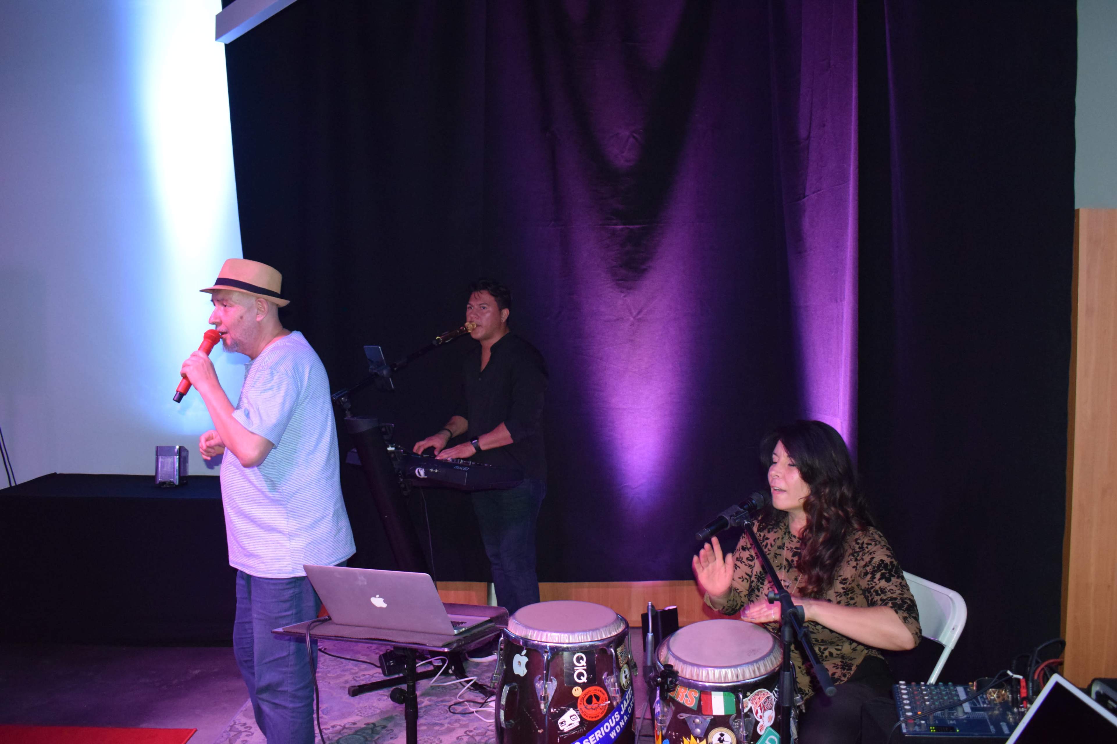 A man sings into a microphone while a woman plays conga drums and a man operates a keyboard behind them, set against a backdrop of purple lighting.