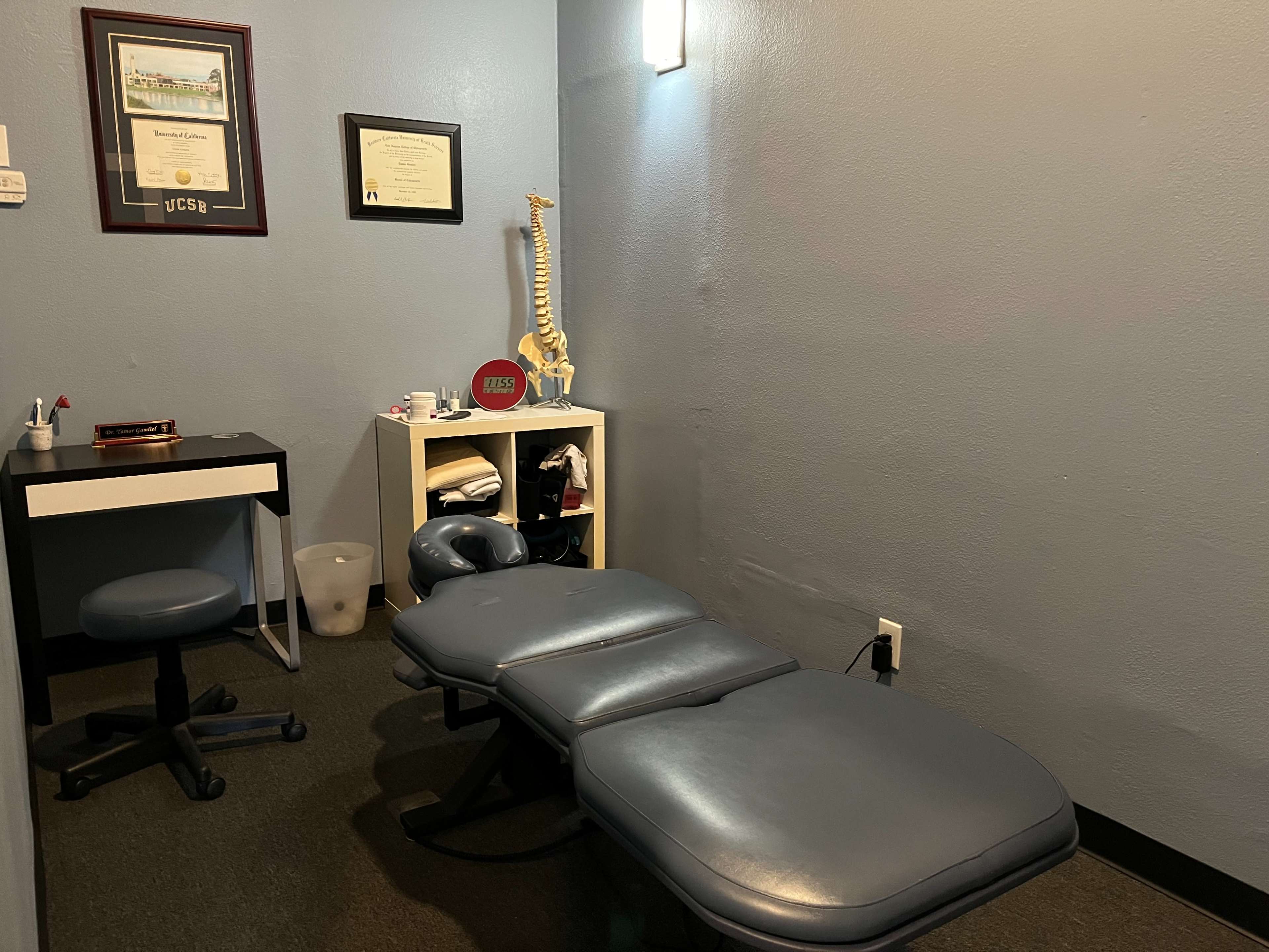 The image shows a small therapy room equipped with a treatment table, a chair, a desk, and a framed certificate on the wall.