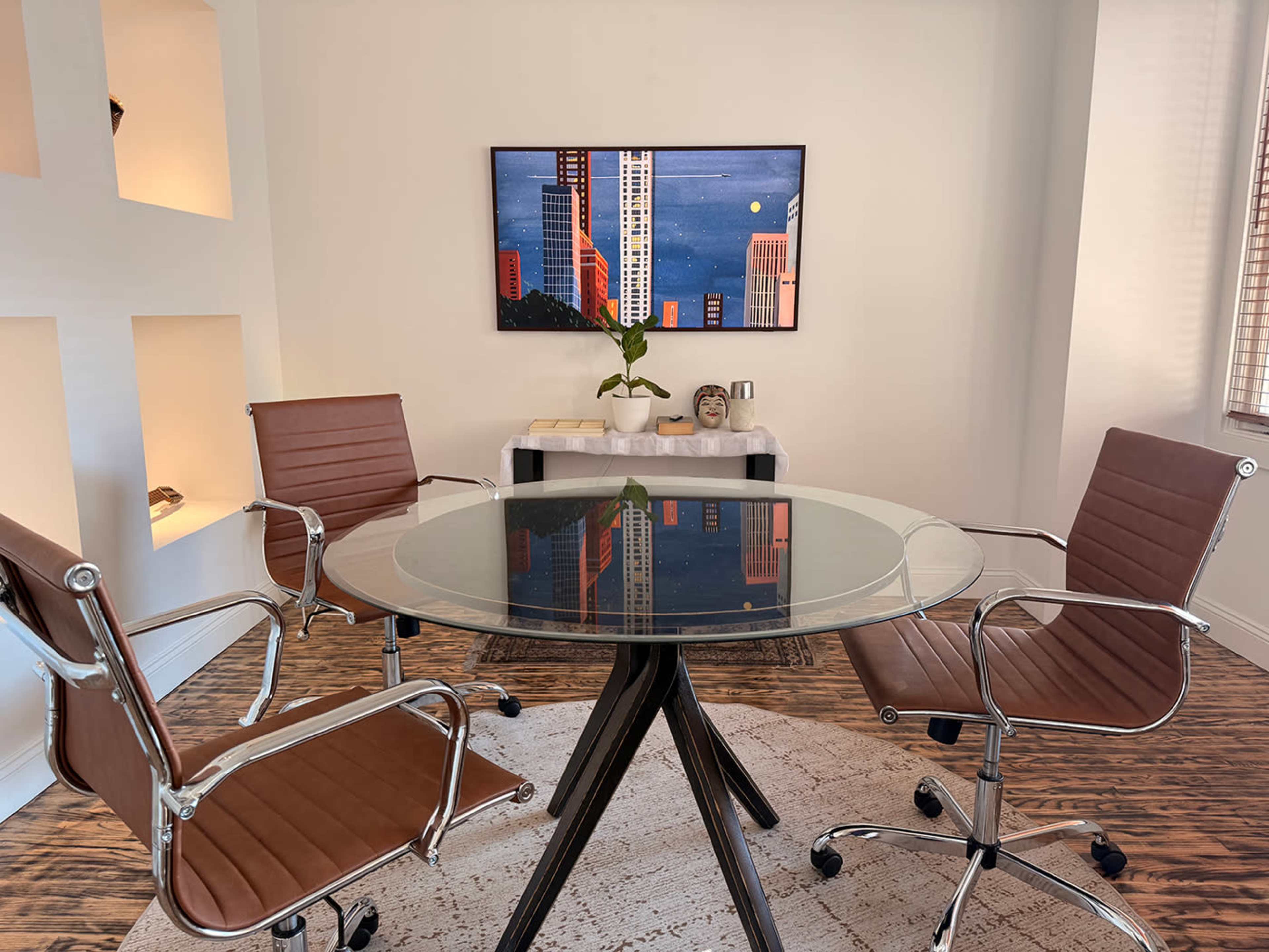 A modern conference room features a circular glass table with four brown leather chairs and a mounted artwork on the wall.