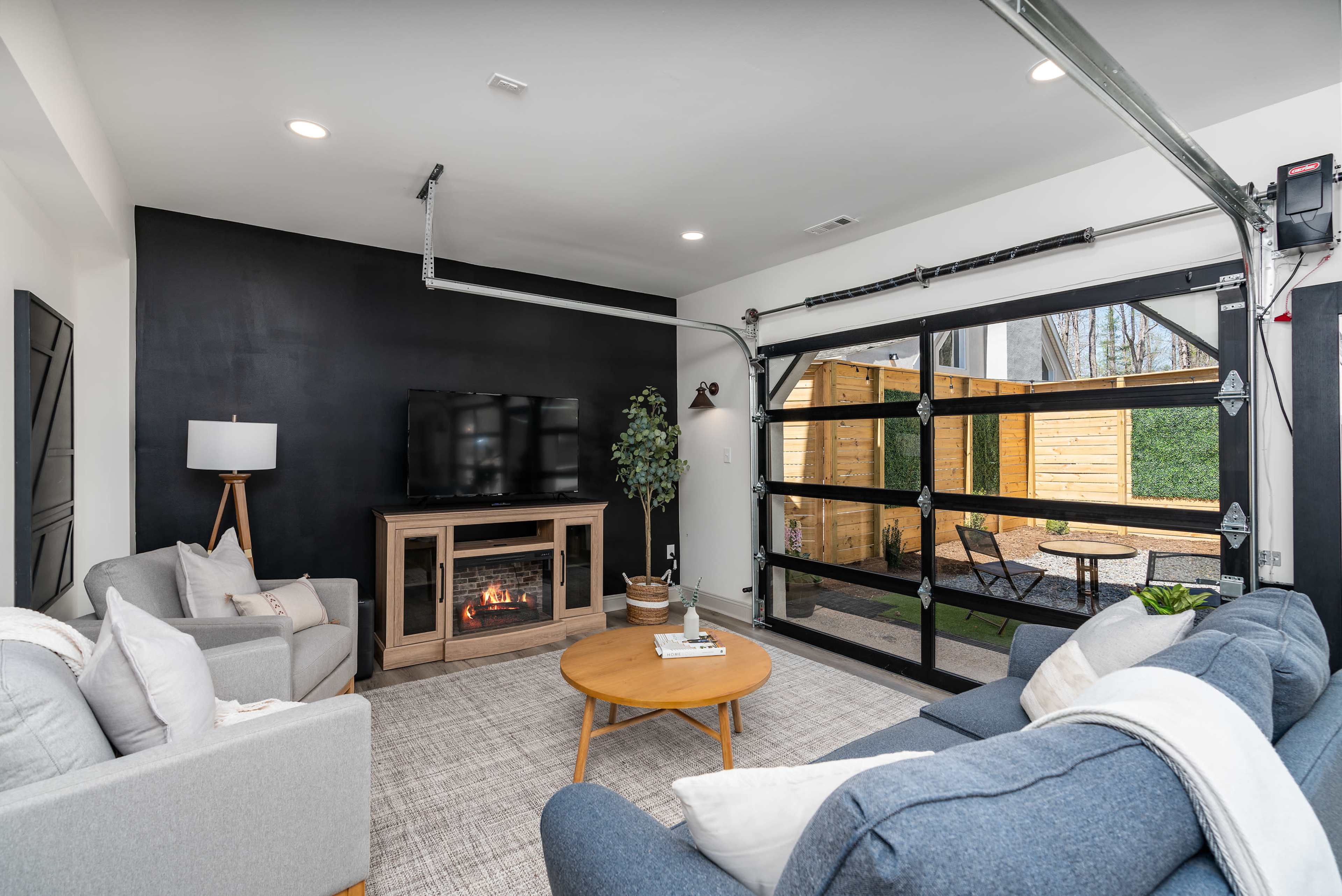 A modern living room with a fireplace, a television mounted on the wall, and a large glass garage door leading to an outdoor patio area.