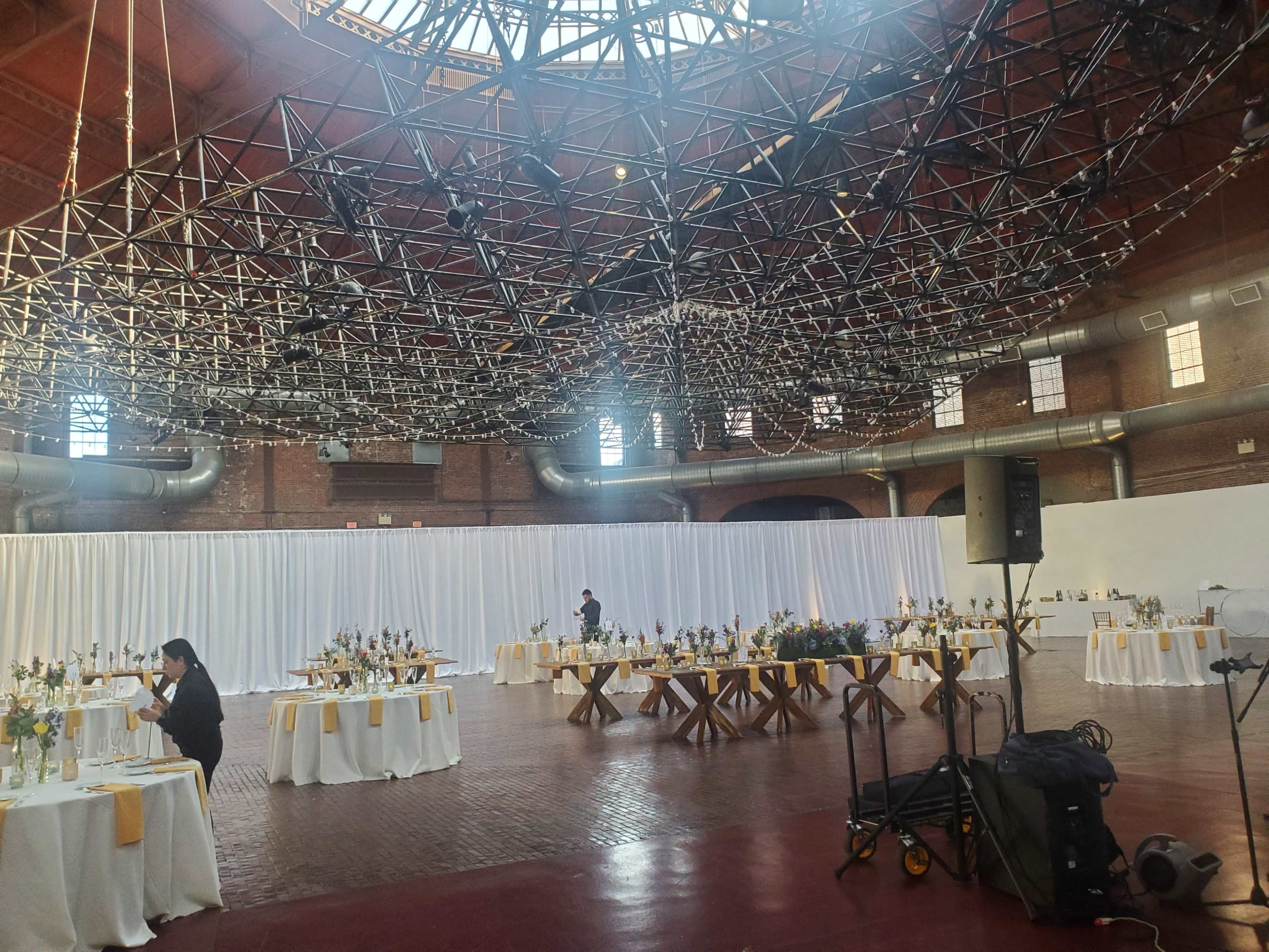 A spacious event hall with decorated tables, a white backdrop, and industrial-style architecture featuring a network of overhead beams.