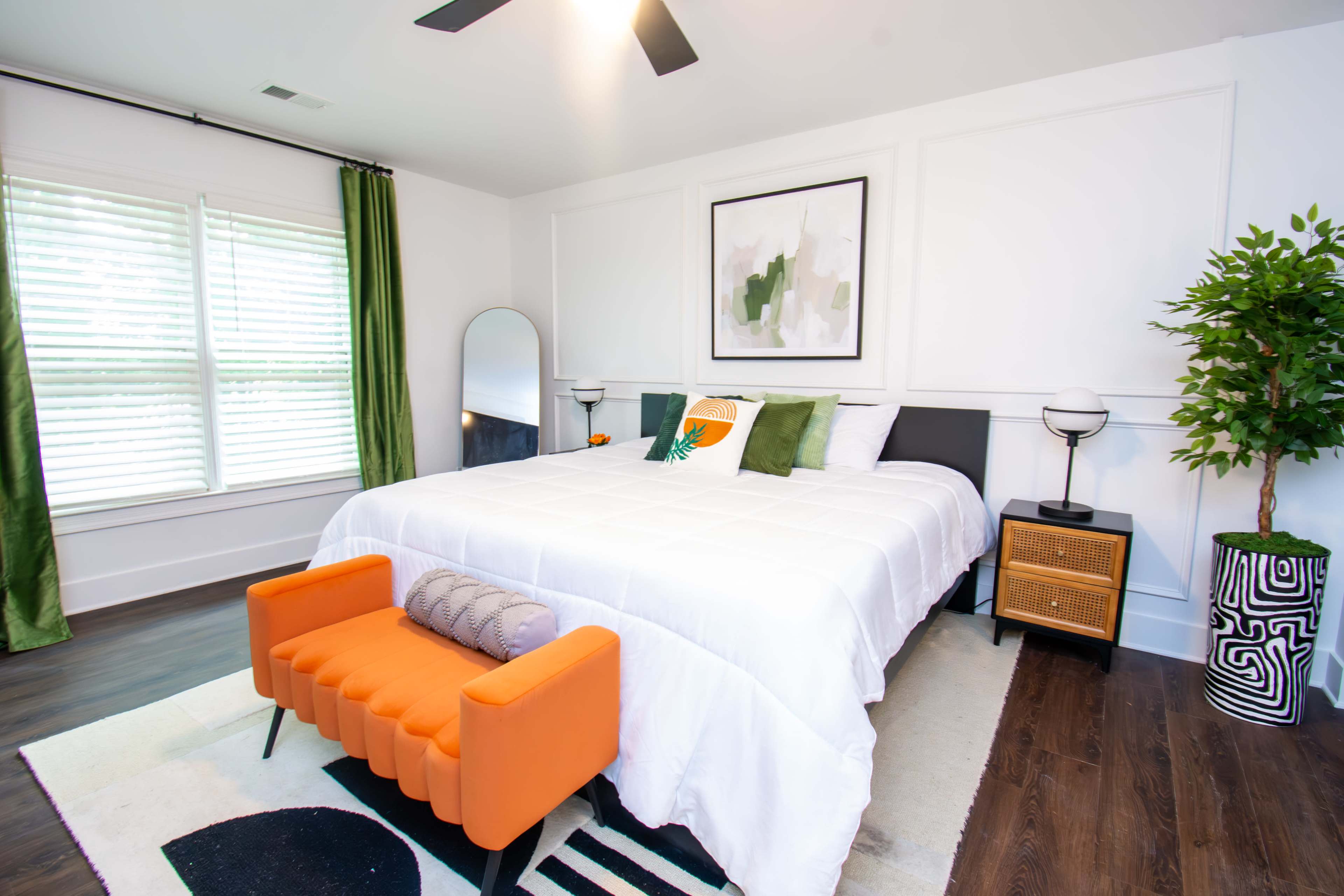The image shows a modern bedroom featuring a large bed with white bedding, an orange accent chair, a green plant, a round mirror, and light green curtains.