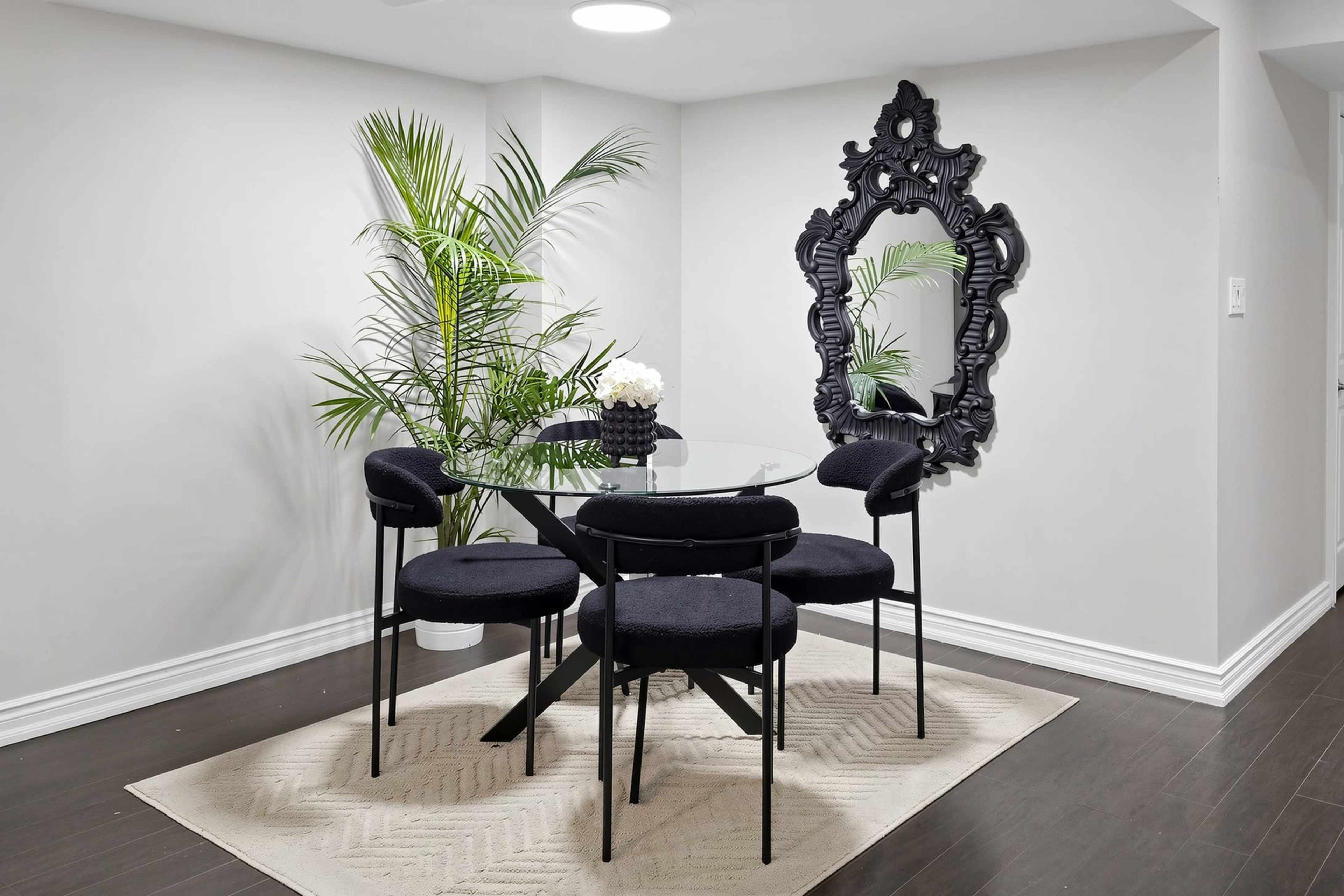 A modern dining area features a round glass table surrounded by six black chairs, with a large ornamental mirror and a potted plant against the wall.