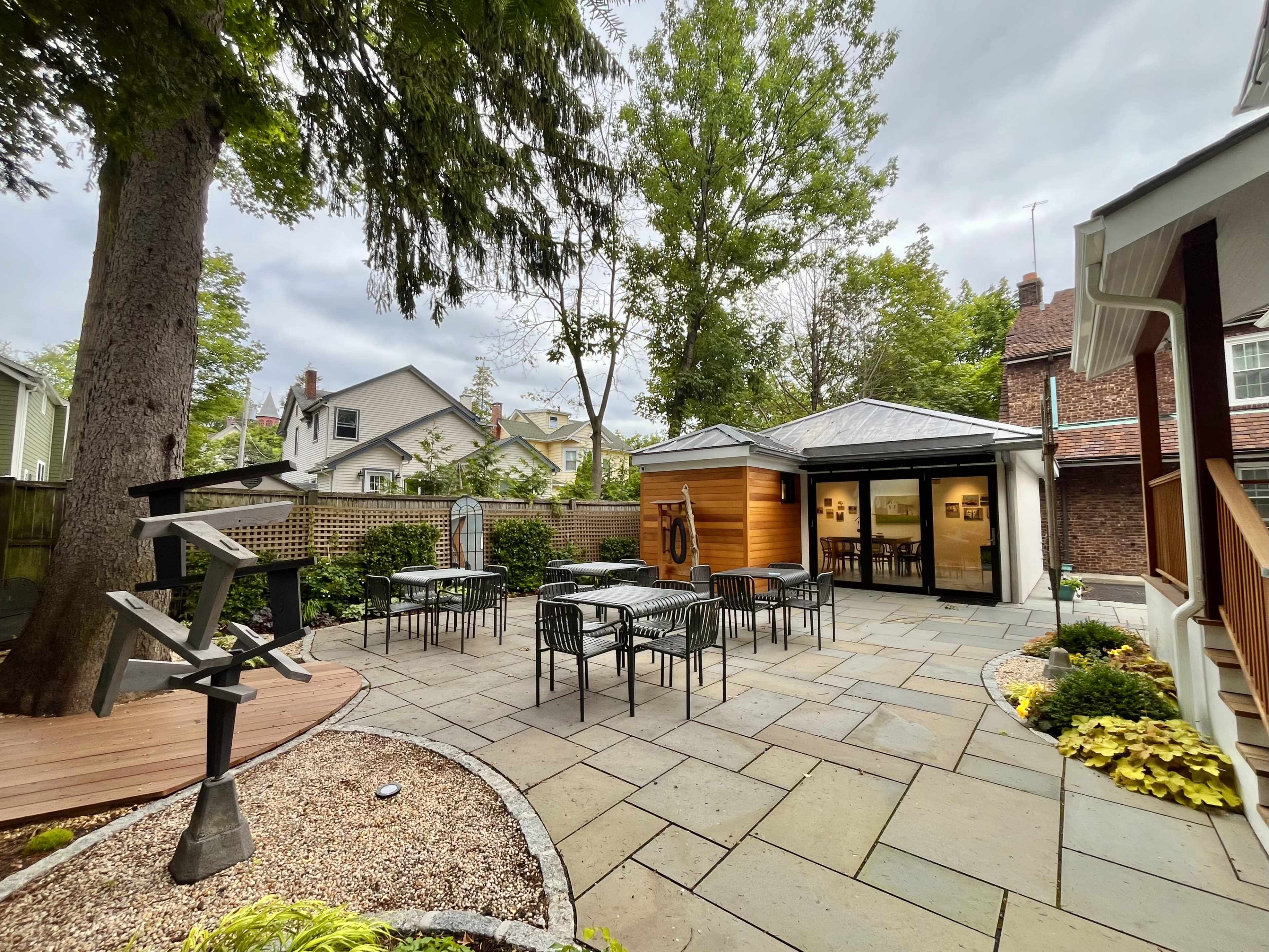 The image shows a landscaped patio area with stone flooring, outdoor tables and chairs, and a small wooden deck beside a nearby tree.