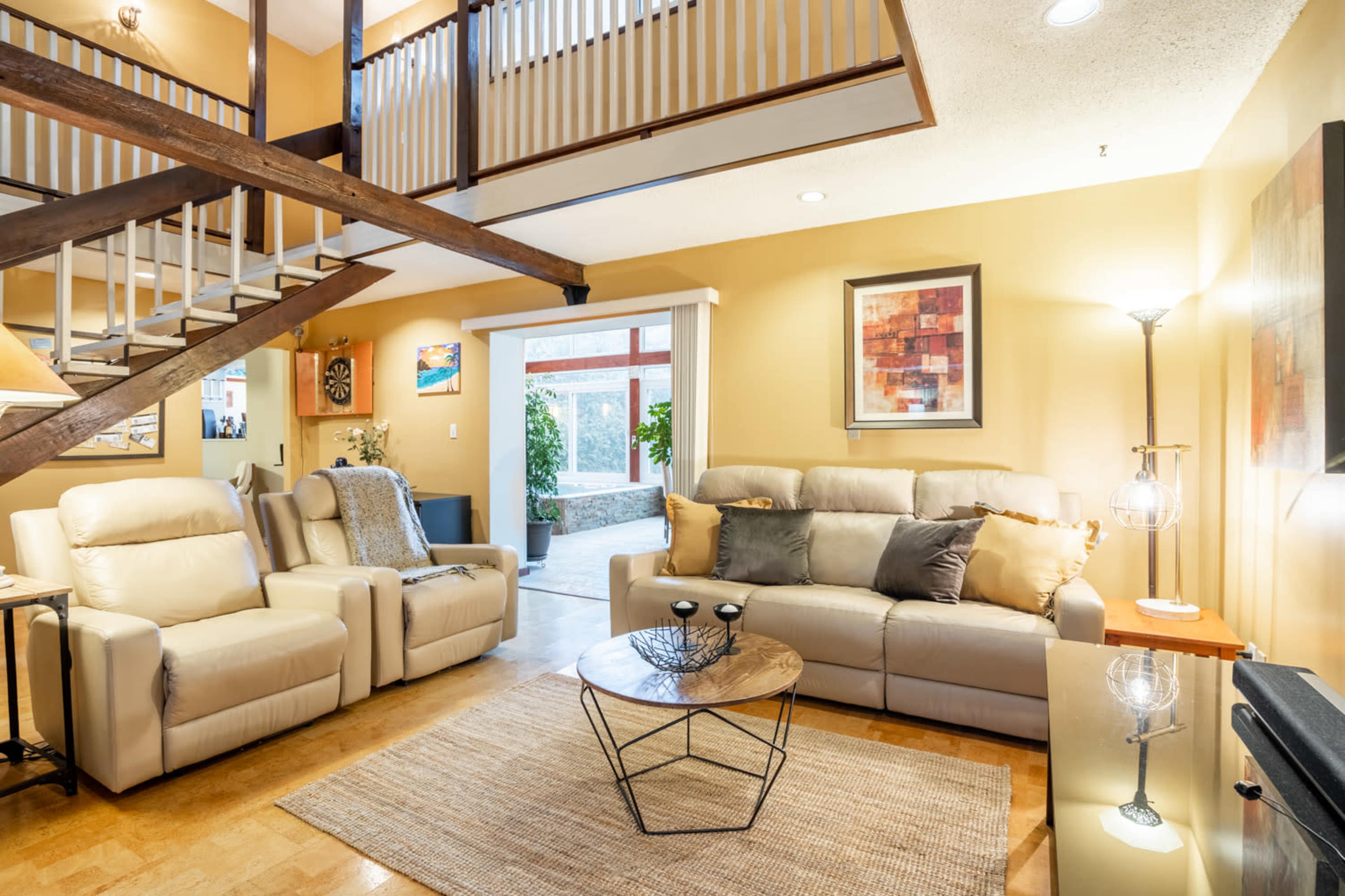 The image shows a cozy living room with beige leather sofas, a glass coffee table, and a staircase leading to an upper level.