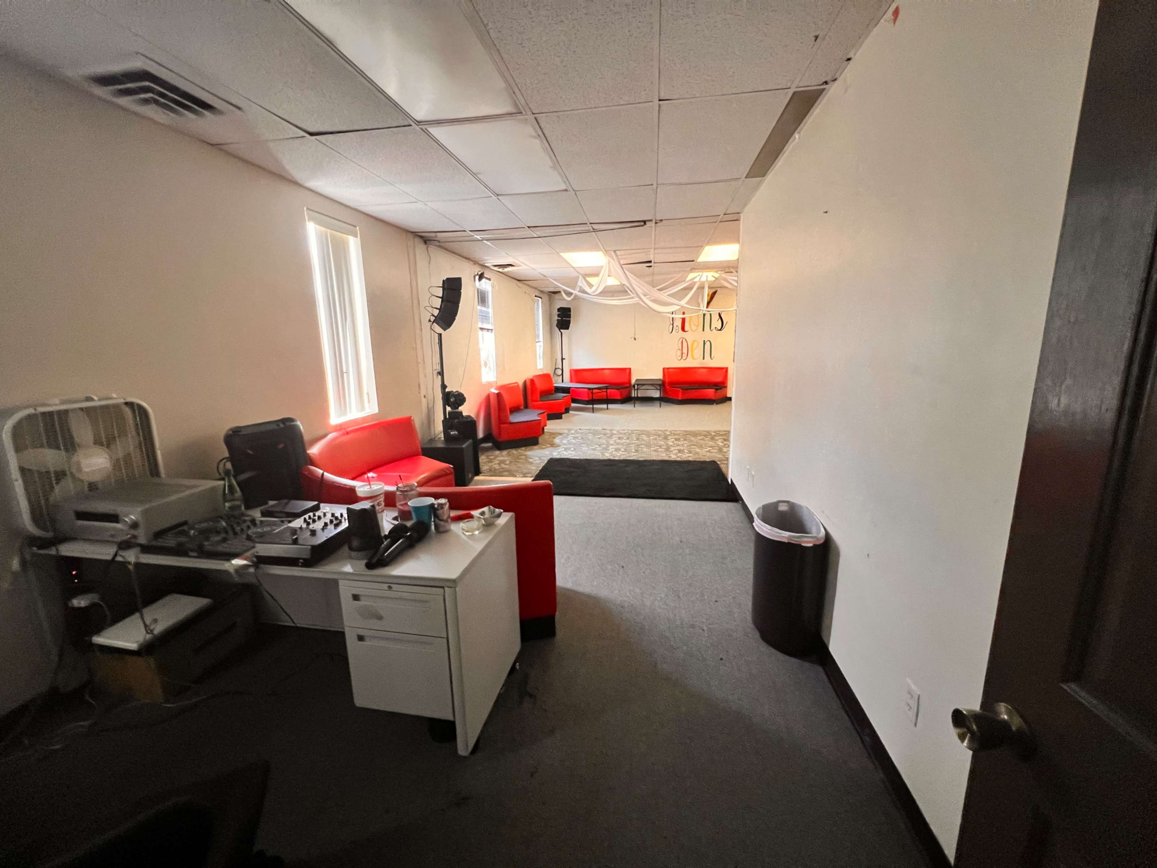 A room with red couches arranged in a seating area, a table with equipment on one side, and a large empty space in the center.