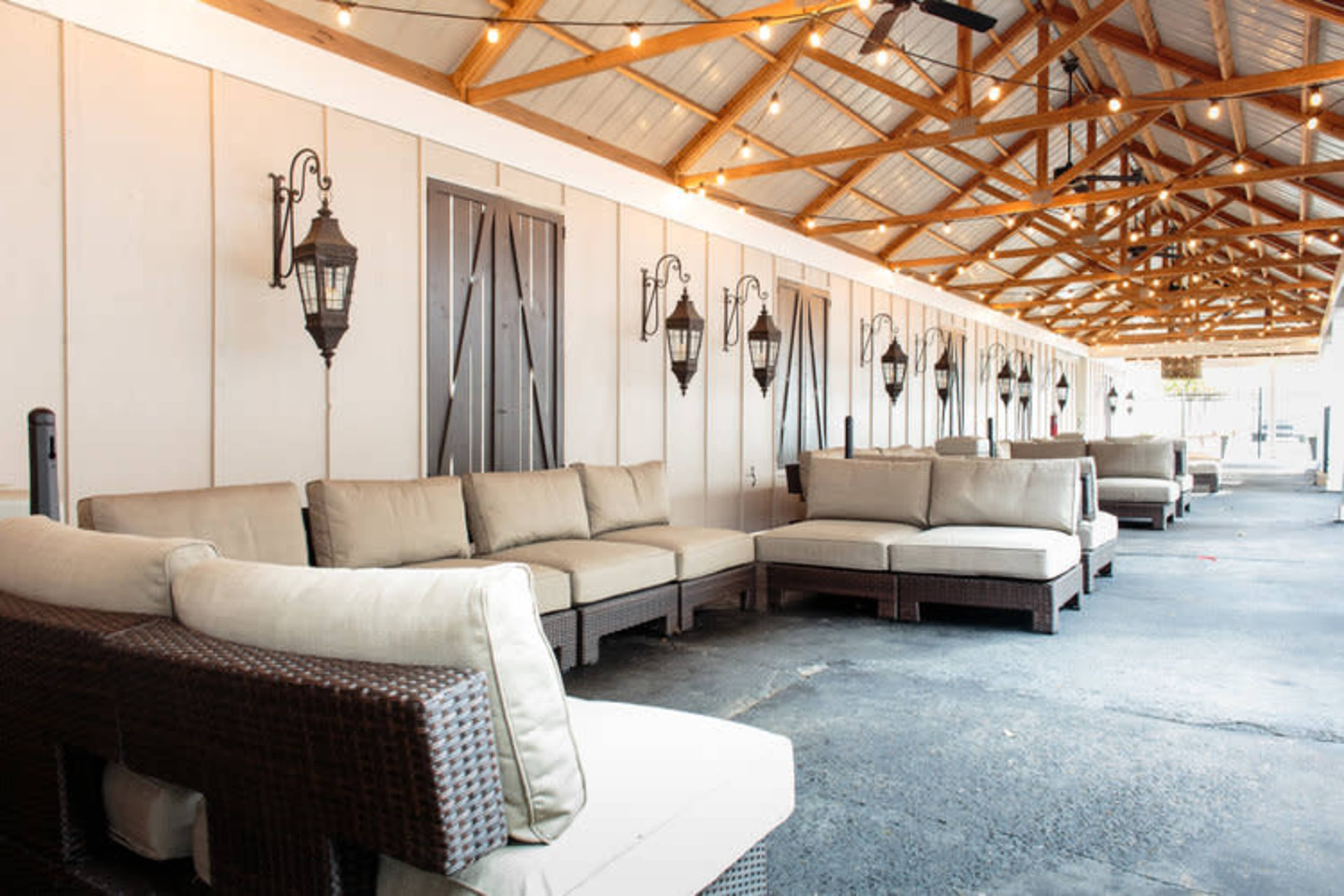 An outdoor lounge area with comfortable couches arranged under a wooden roof, illuminated by string lights and featuring wall-mounted lanterns.